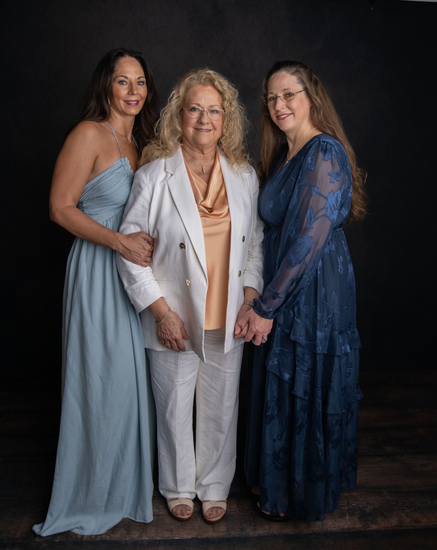 Three women in contrasting dresses pose together in a series of intimate portrait shots against a dark background.