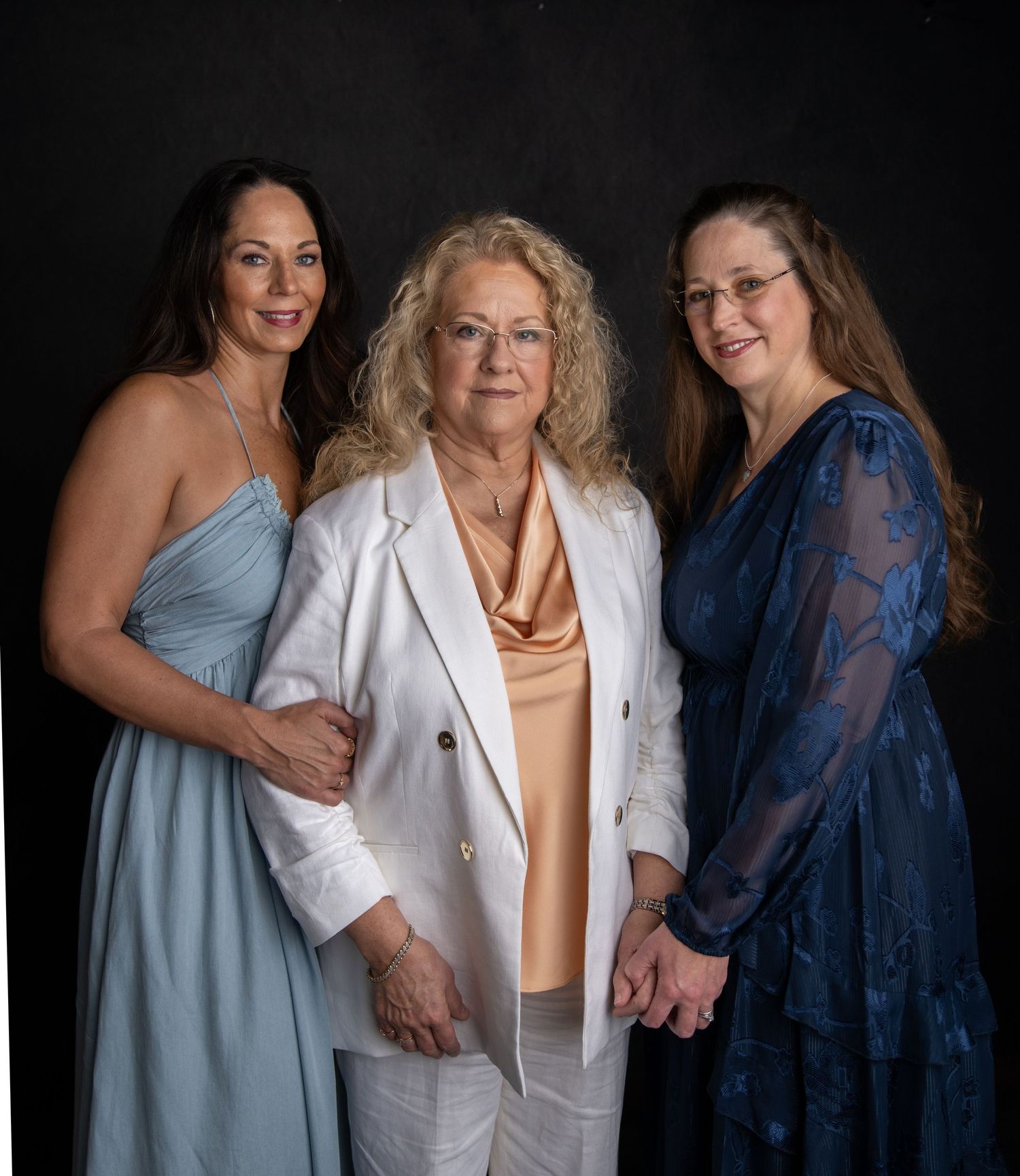 Three women pose together against a dark background wearing blue and white outfits in a professional studio setting.