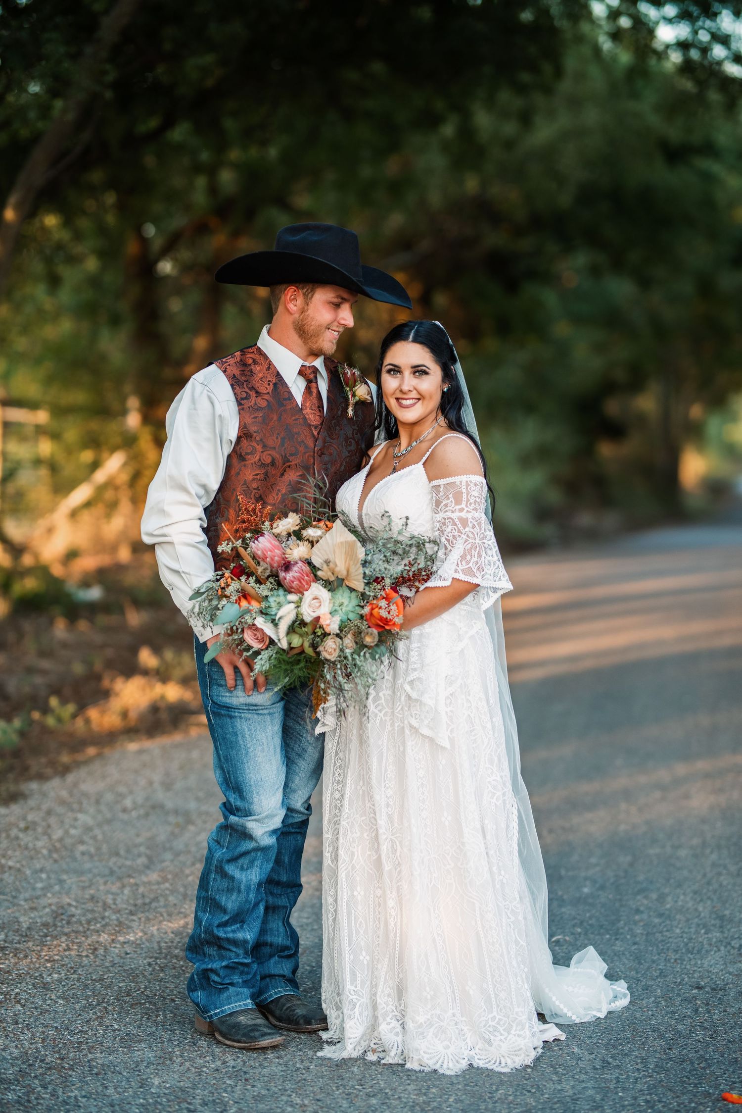 Romantic western-themed couple in wedding attire with floral bouquet on country road.