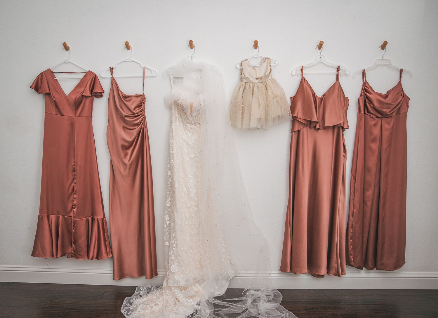Wedding party dresses in coral and white displayed on hangers against a gray wall.