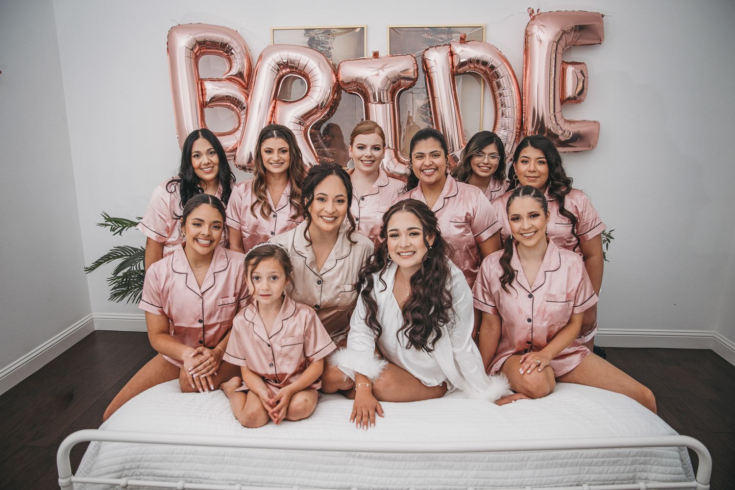 A bridal party wearing matching pink satin robes poses together on a bed under rose gold letter balloons spelling BRIDE.