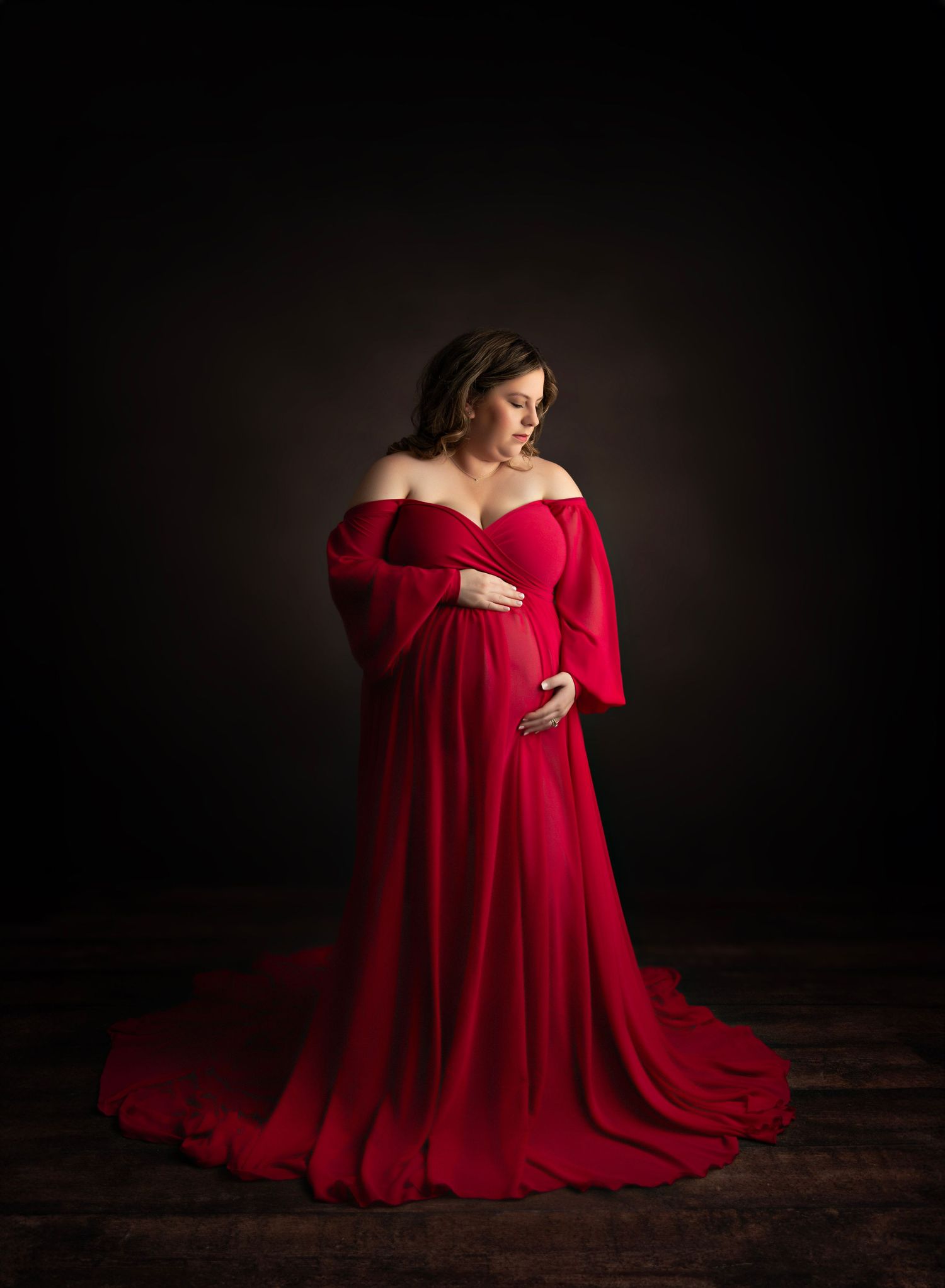 Dramatic maternity portrait featuring woman in red off-shoulder gown with long train.
