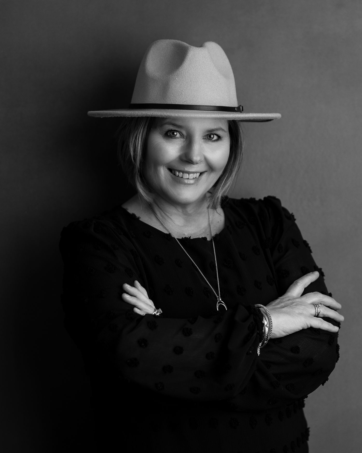 Black and white portrait of a person wearing a wide-brimmed hat and black sweater, standing with arms crossed and smiling.