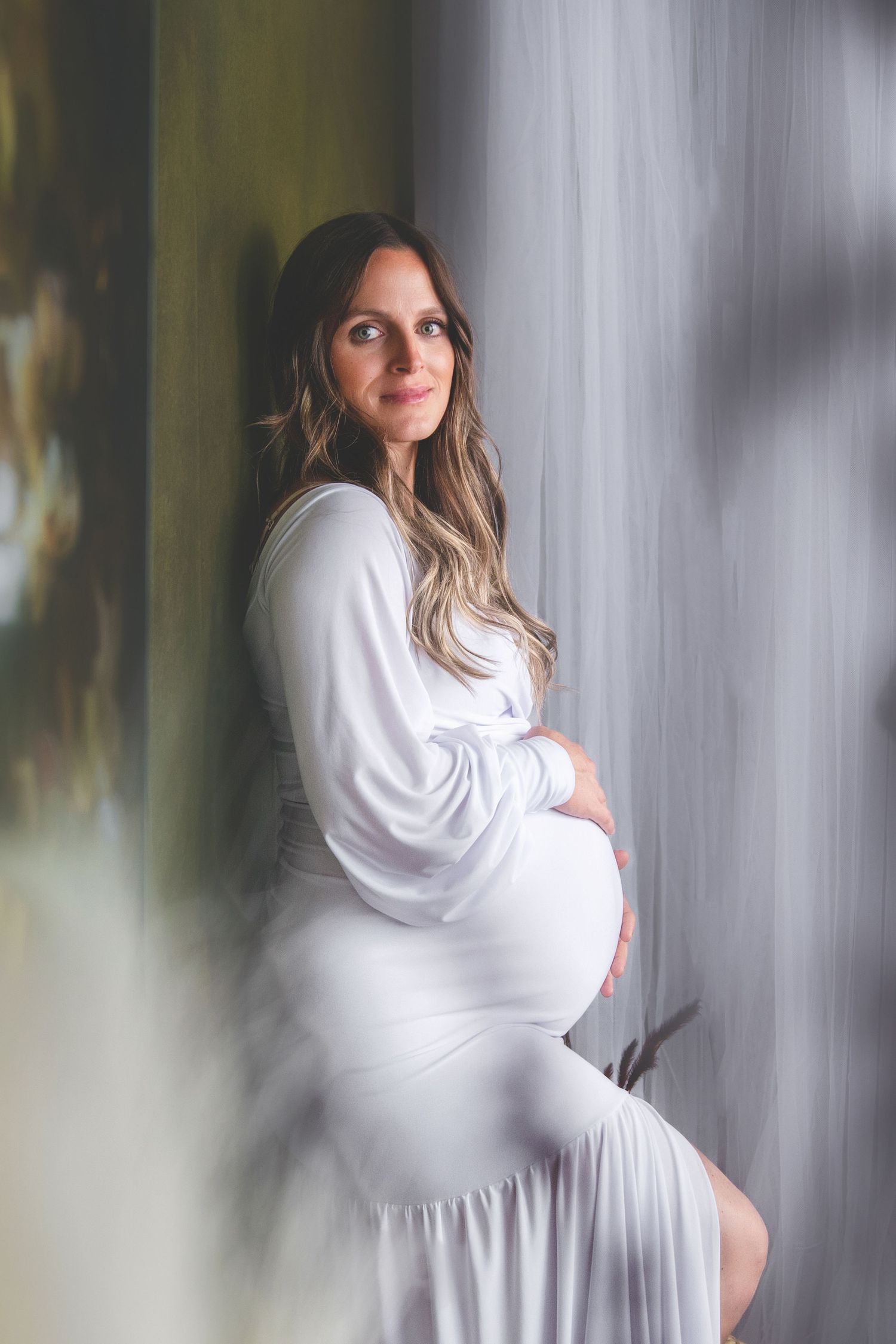A dreamy maternity portrait features an expectant mother in a flowing white dress posing against a soft, ethereal background.