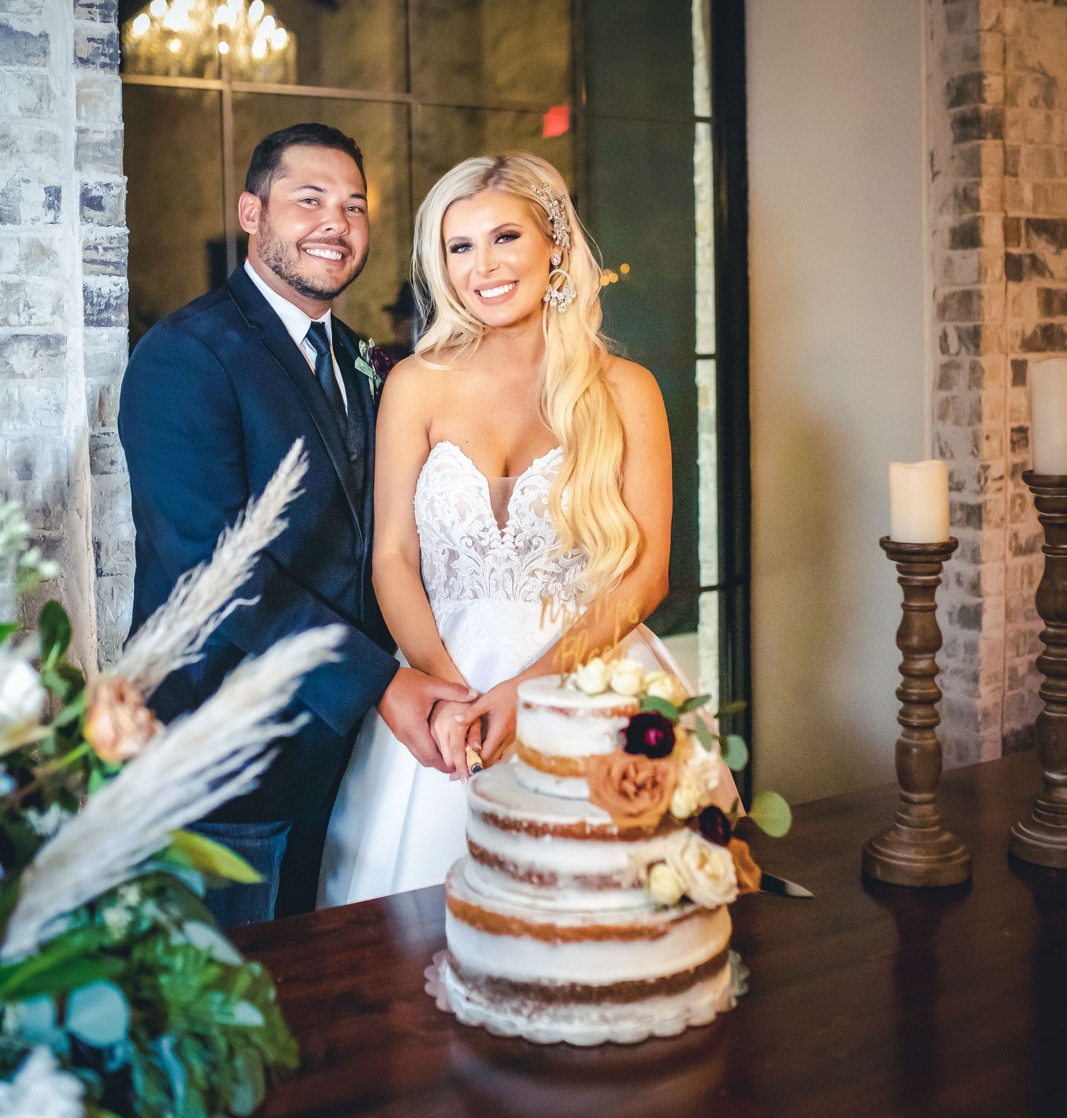 A couple poses by their rustic naked wedding cake decorated with fresh flowers.