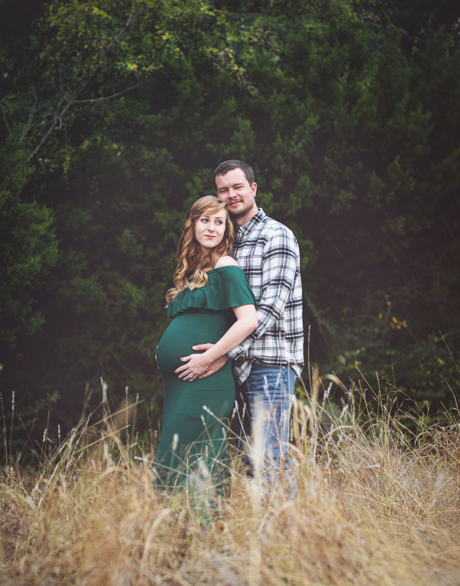 Couple poses together in tall grass with forest backdrop during maternity photoshoot.