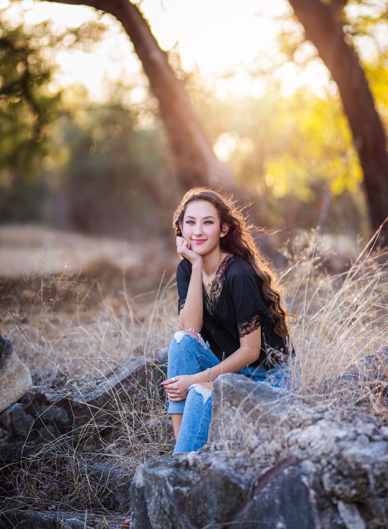 Young person in casual wear poses on rock with sunlit trees in background.