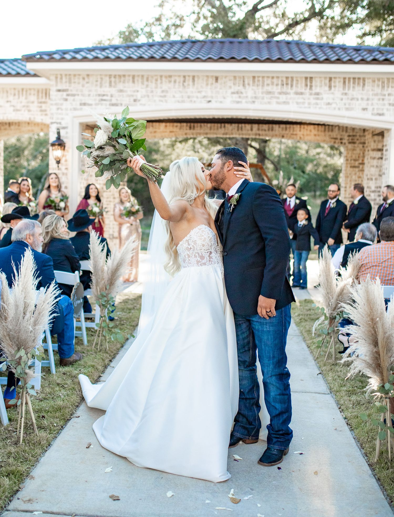 A romantic first kiss at a wedding ceremony with pampas grass decor and stone architecture.