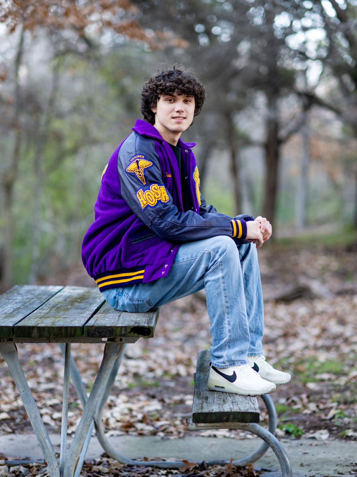Person wearing purple letterman jacket sits casually on picnic table in autumn woods.
