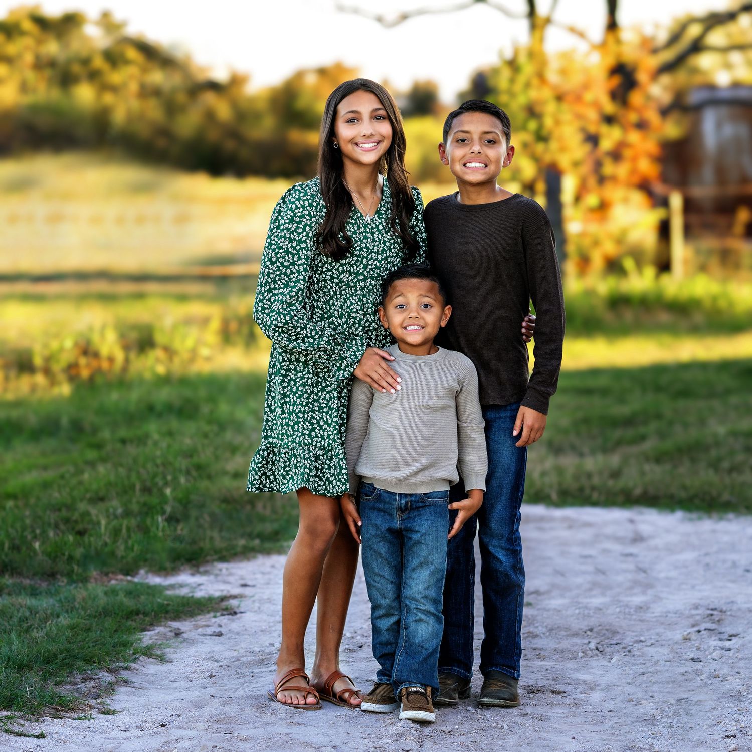 Family of three poses together on dirt path with autumn foliage in background.