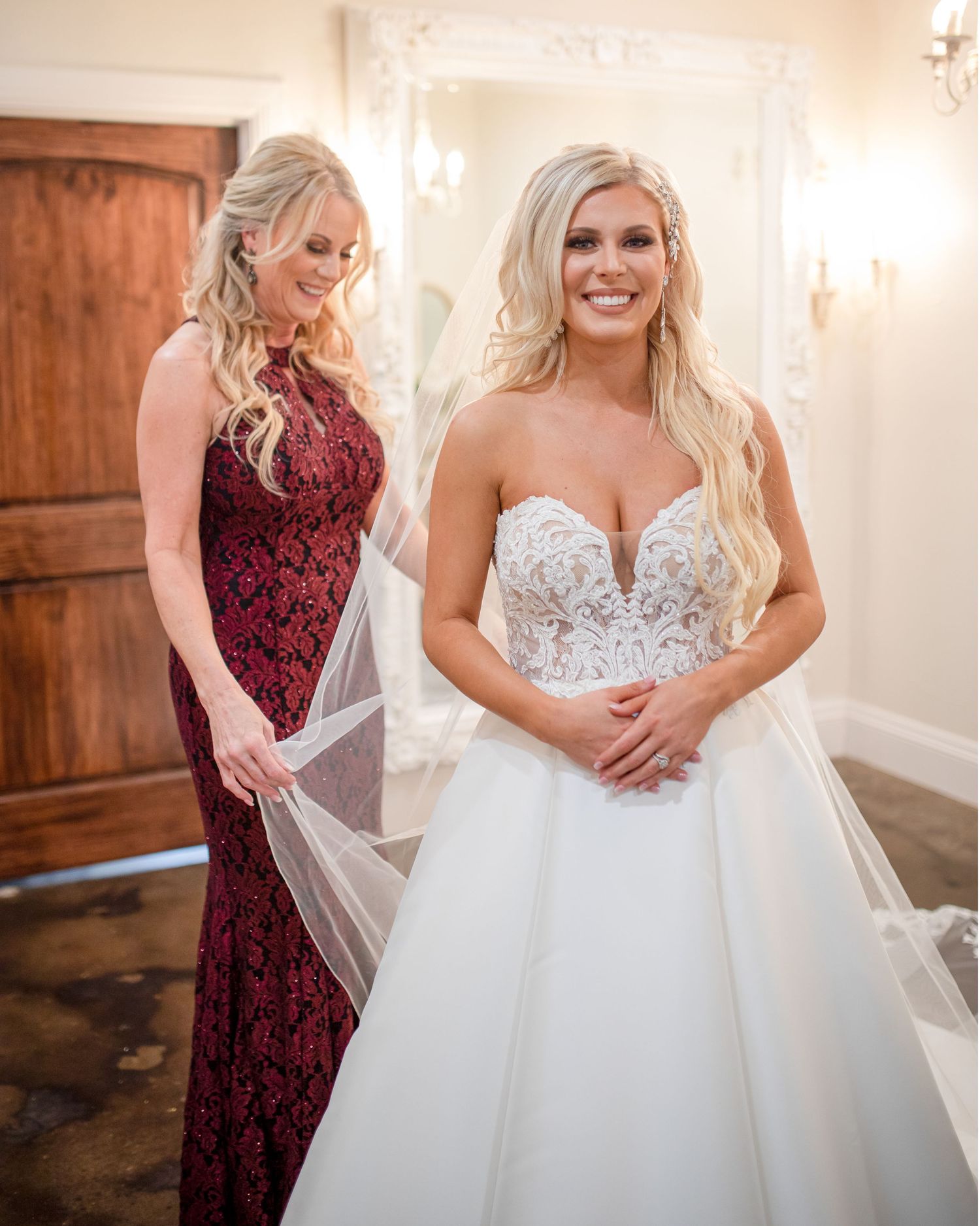 Two women in formal gowns share a joyful moment in an elegant dressing room.