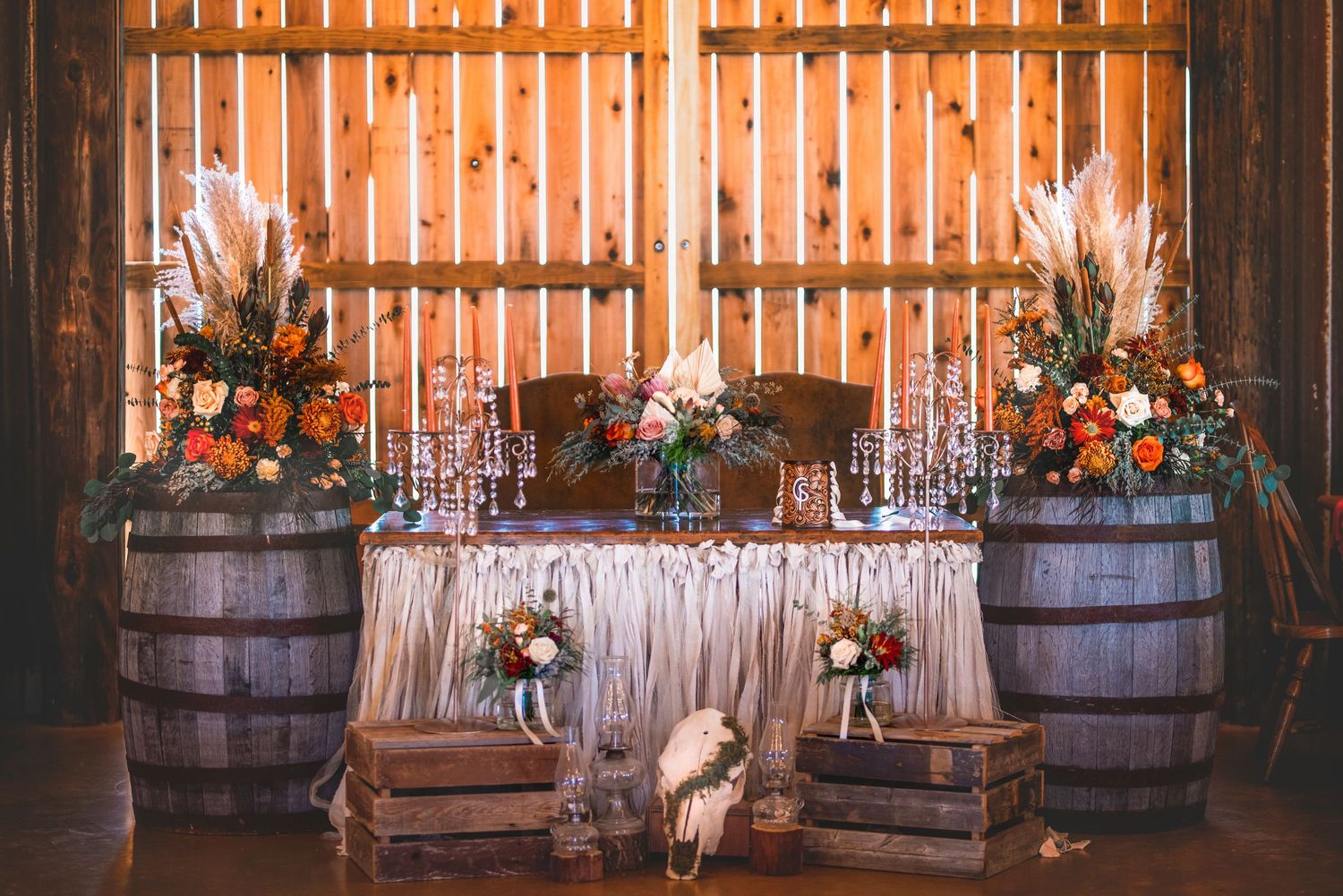 Rustic wedding reception setup with wine barrels, wooden crates, and floral arrangements against wooden wall.