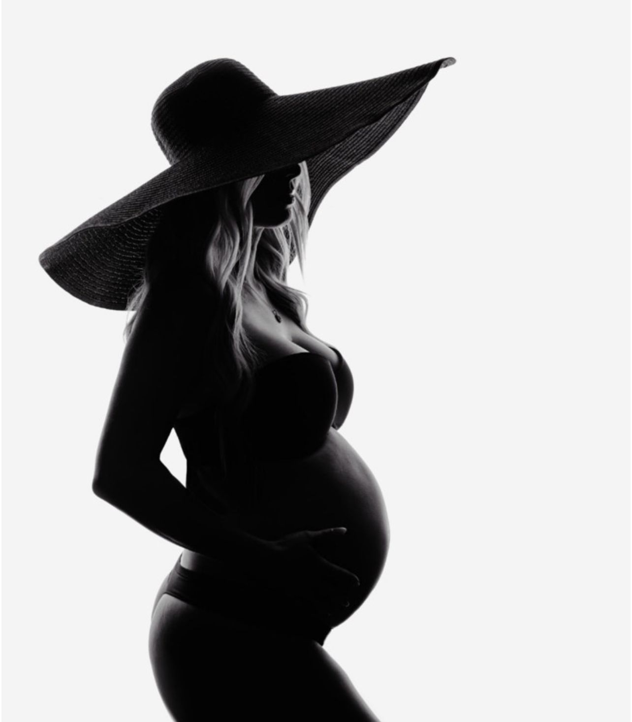 Artistic silhouette maternity portraits showing pregnant woman in wide brim hat against white background.