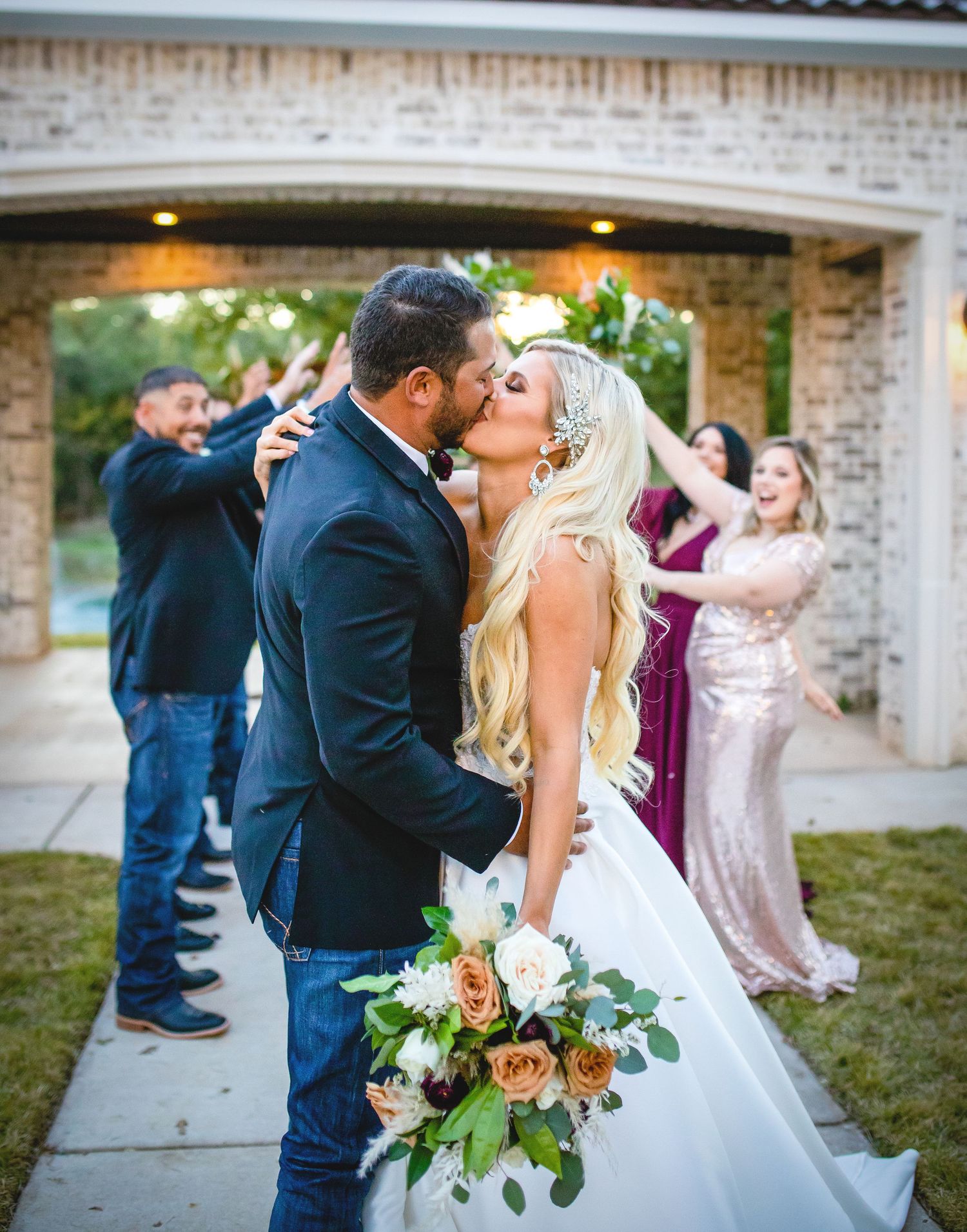 A couple shares their first kiss as guests celebrate in the background near a brick archway.