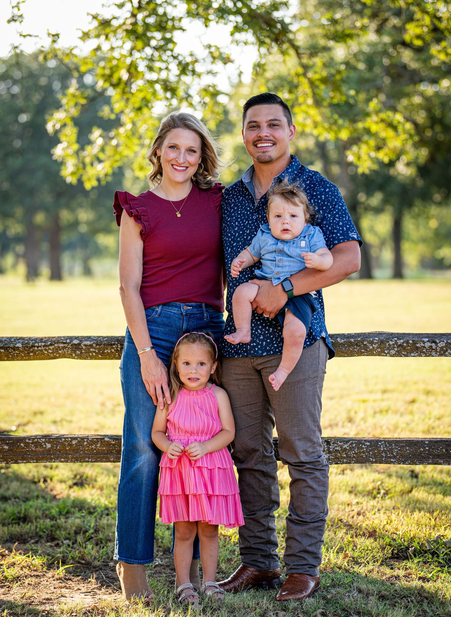A family poses together by a wooden fence during golden hour in a grassy field on a sunny autumn day.