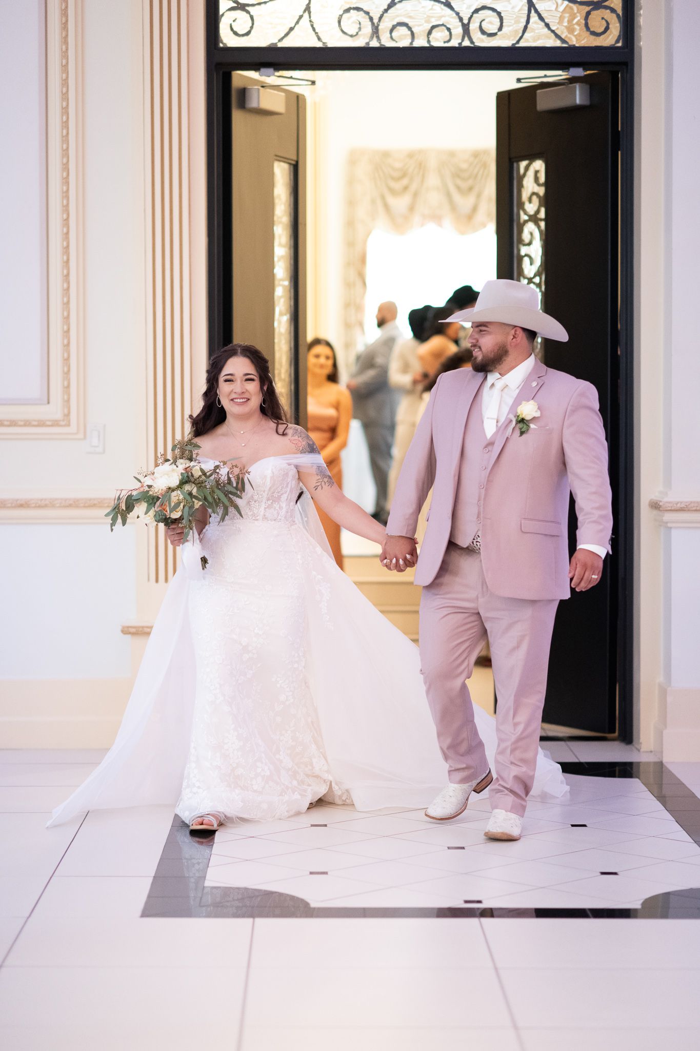 A newlywed couple exits an elegant doorway in coordinated pink and white wedding attire.