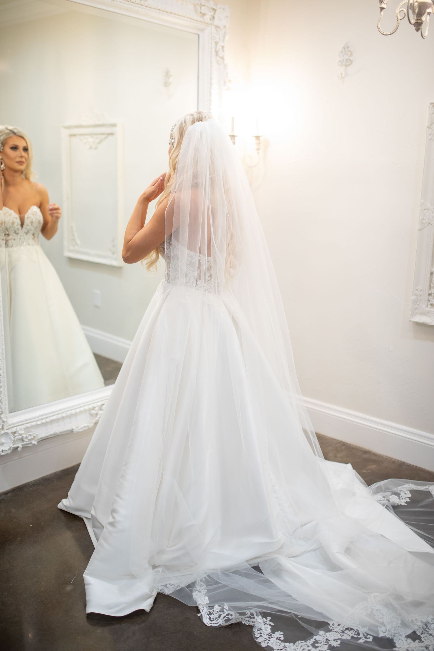 A bride in a white ballgown wedding dress looks at her reflection in an ornate mirror while adjusting her veil.