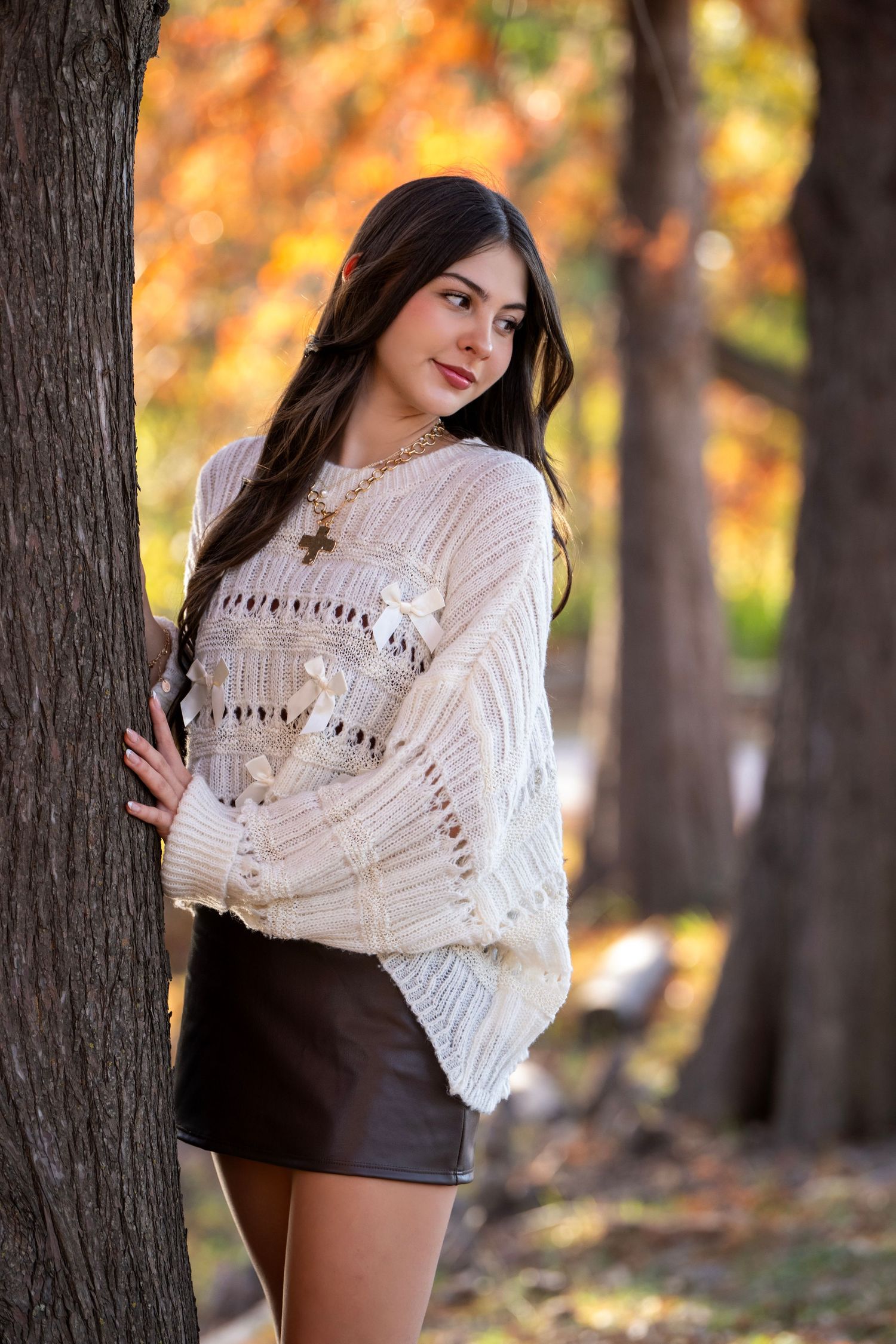 A woman in a white knit sweater and dark shorts poses against autumn trees with golden foliage in the background.