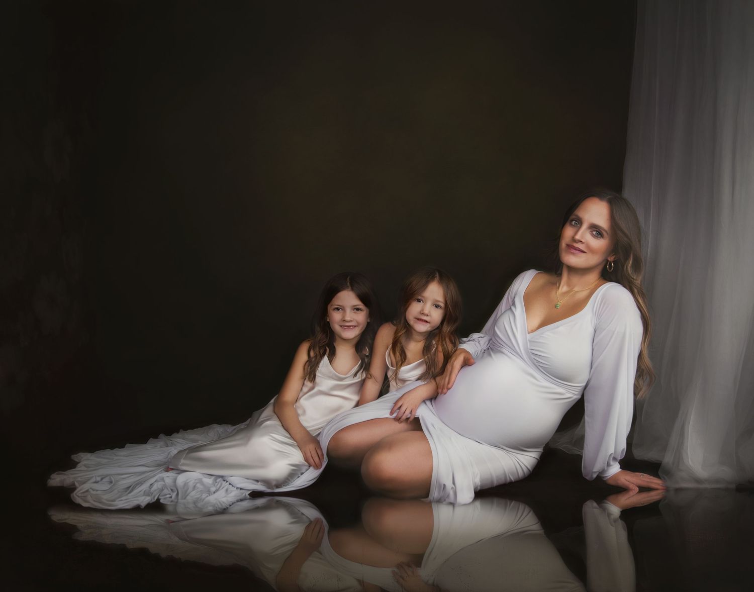 Maternity photo showing three people in white dresses sitting together on the floor in a dark room.