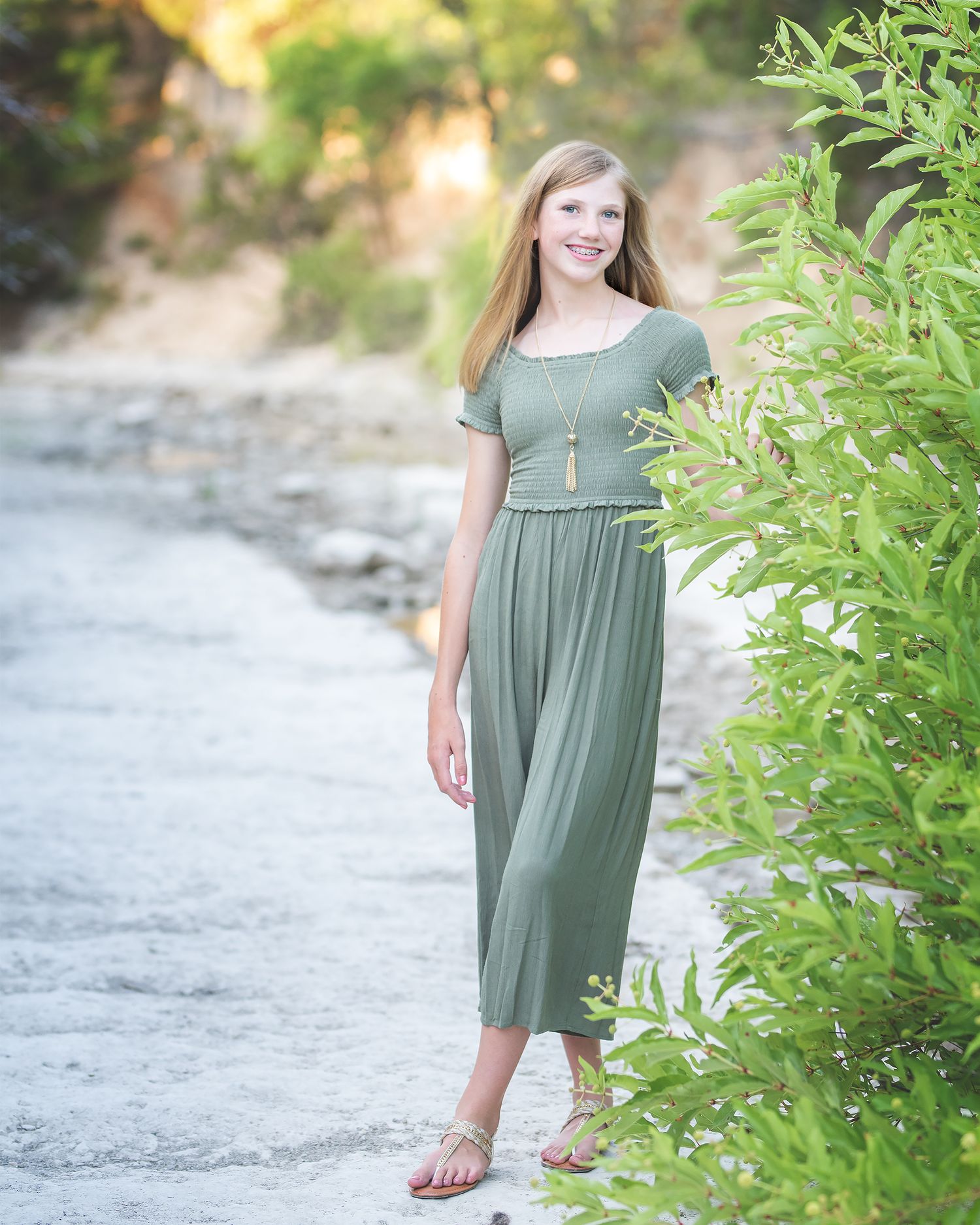 Young person in olive green dress poses outdoors against natural greenery during golden hour sunset.