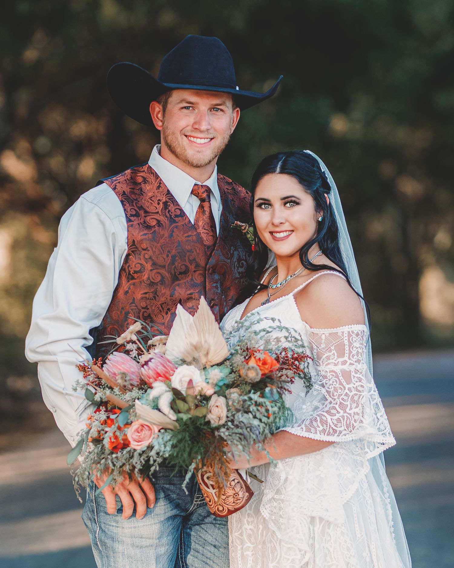 Western-style wedding couple poses together with coral and blush floral bouquet outdoors.