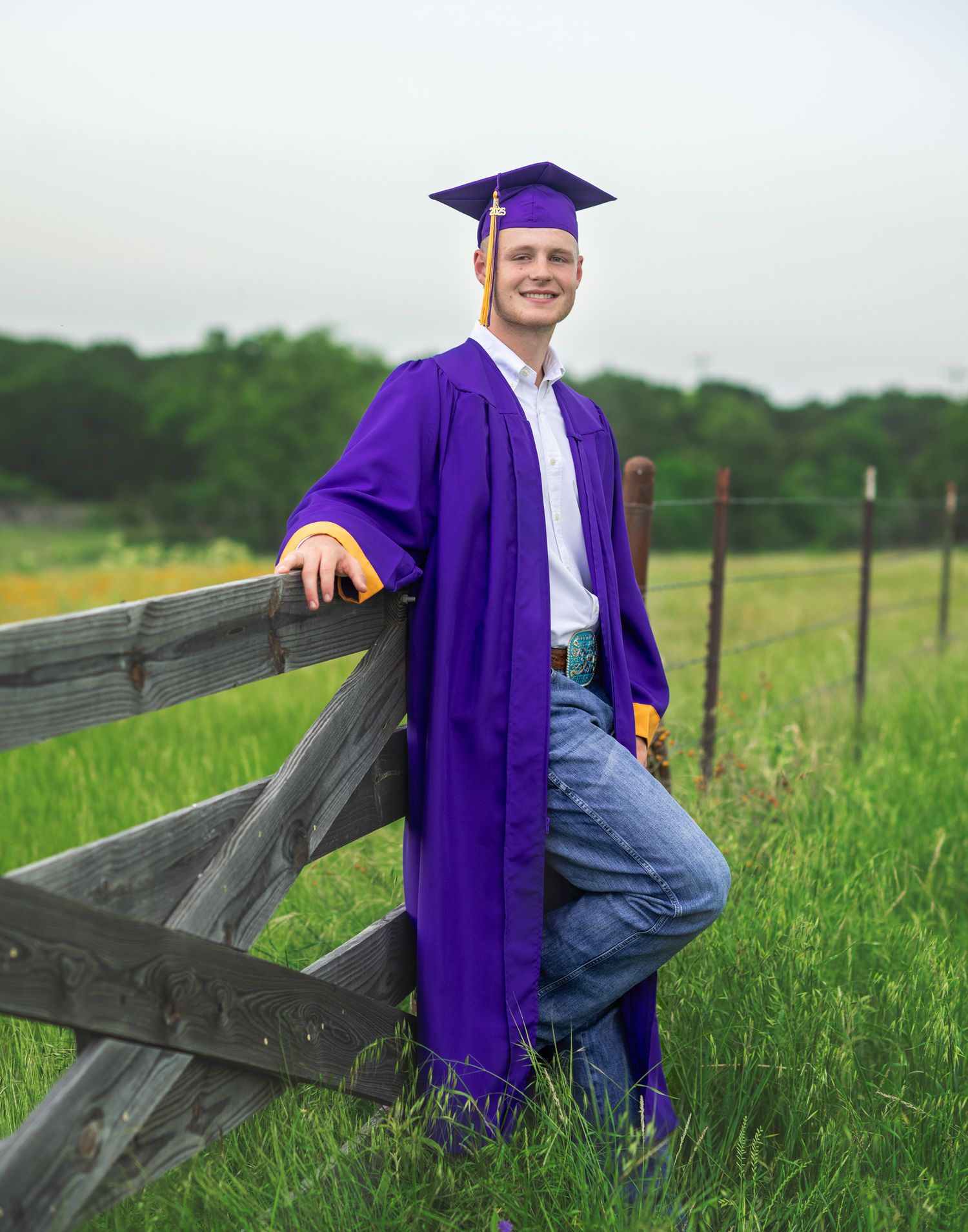 Graduate in purple cap and gown poses casually on wooden fence in green field.