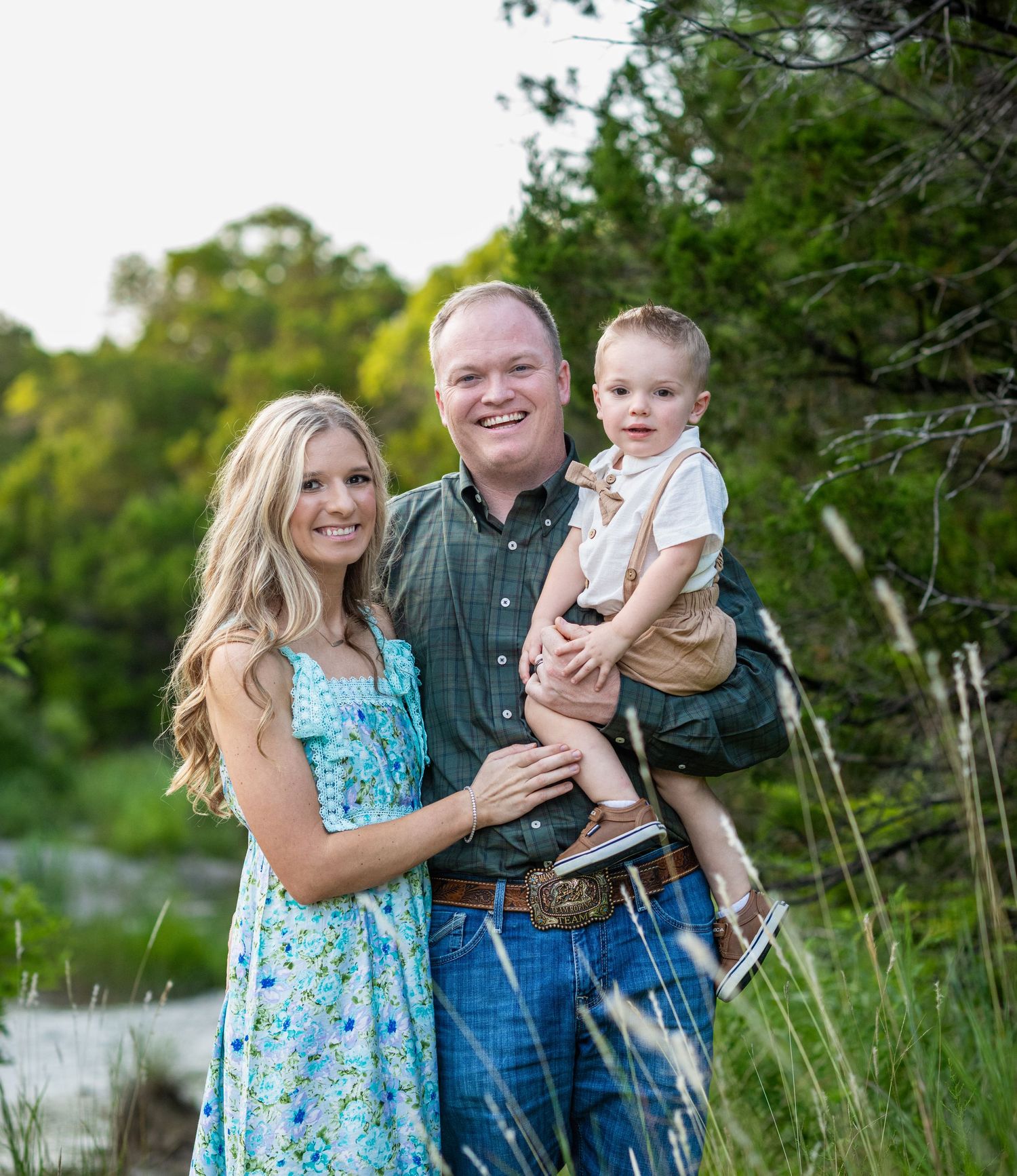 Family of three poses together outdoors in casual clothing against natural greenery.