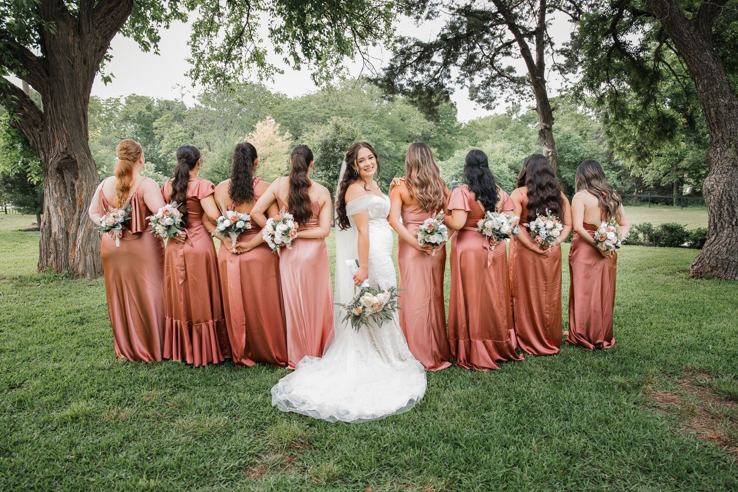 A bridal party photo with bridesmaids in coral dresses standing on grass under trees.