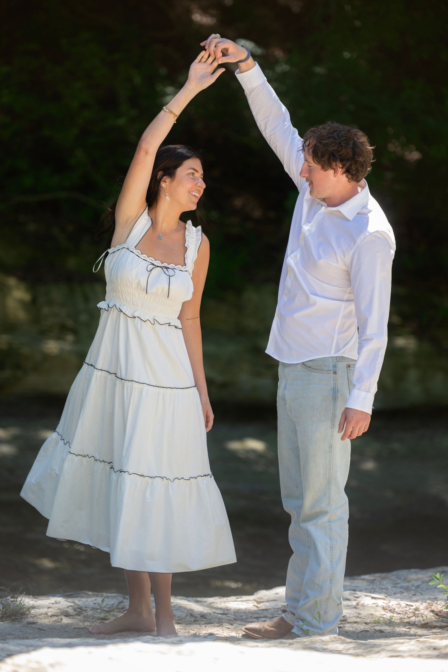 Couple in white outfits dance together on beach in romantic engagement photo.