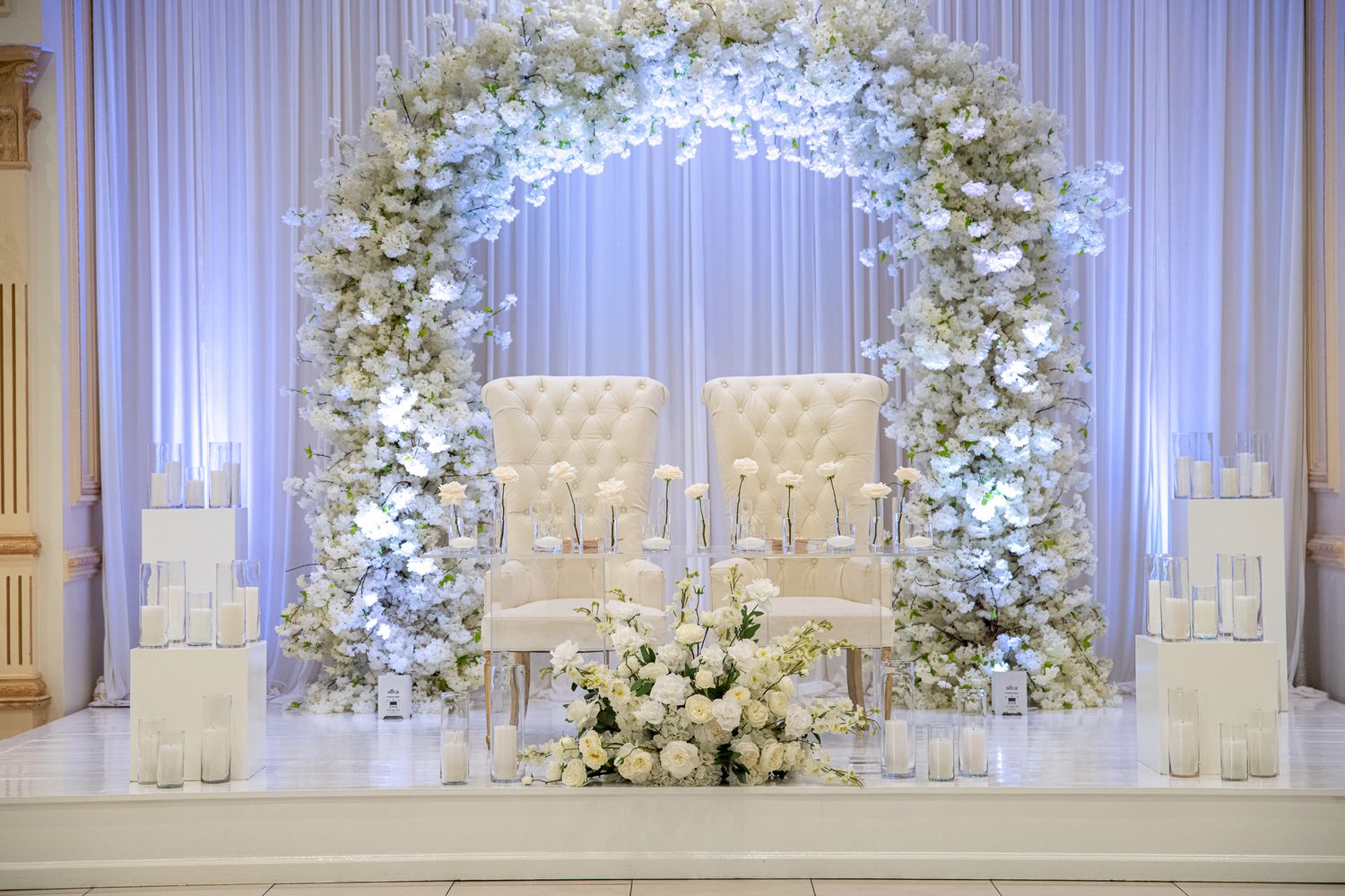 Elegant white floral arch with crystal chandelier and white chairs for wedding ceremony setup.