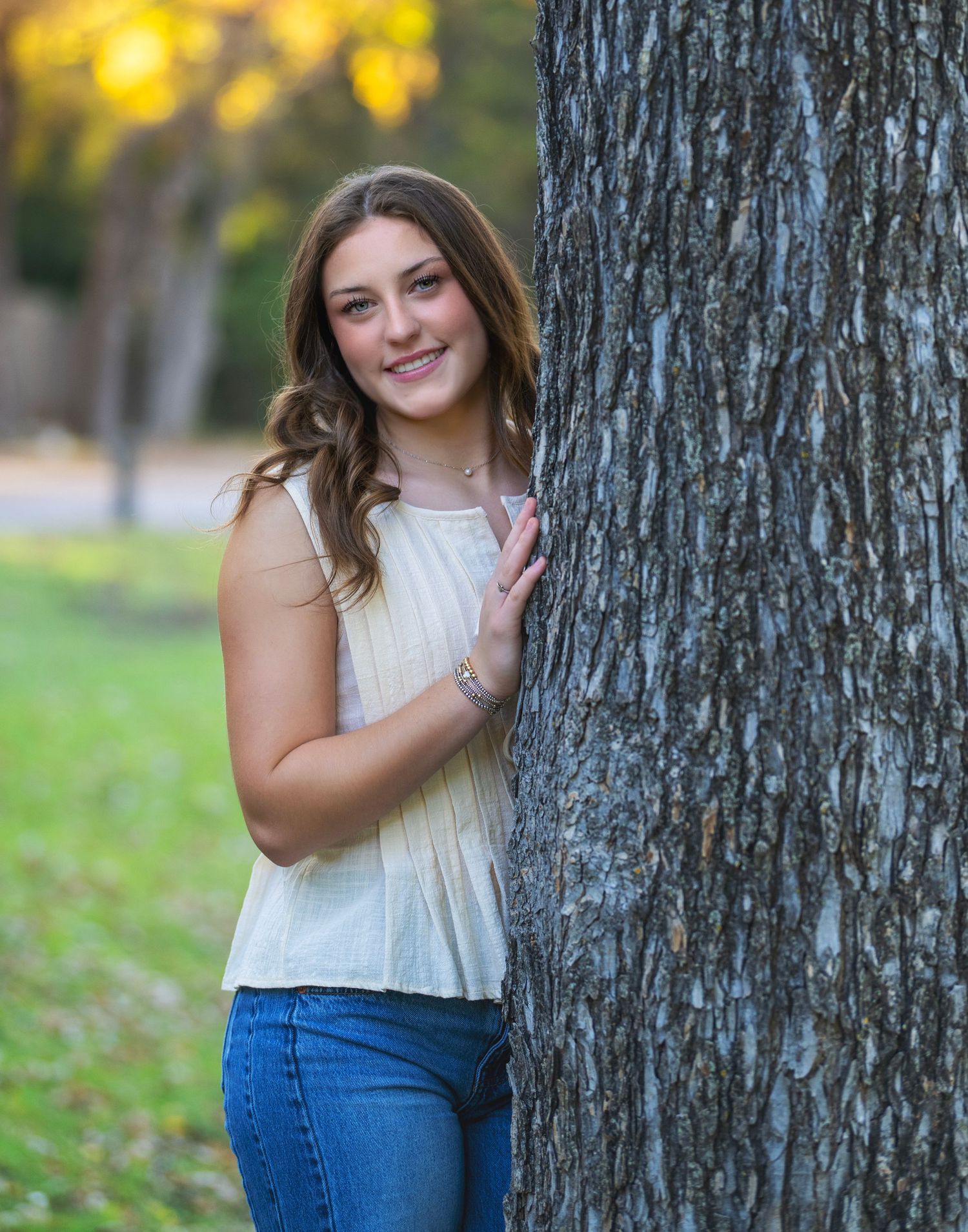 A casual outdoor photoshoot featuring someone in a white sleeveless top and blue jeans posing against a tree trunk.