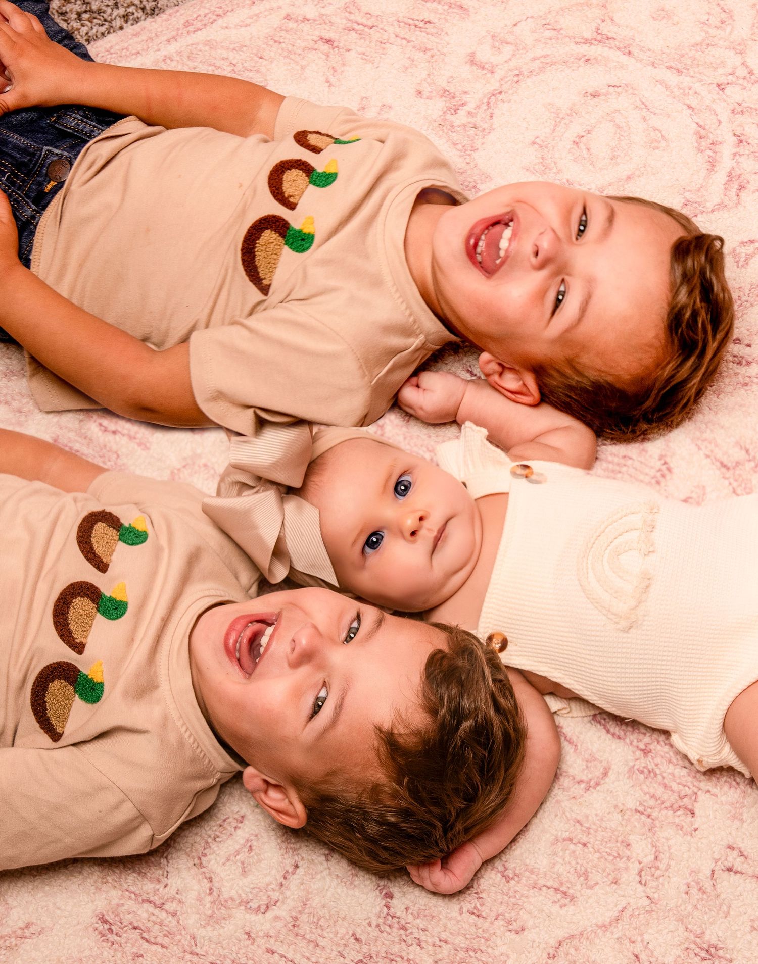Three people laying on pink fabric wearing matching tan shirts with avocado designs.