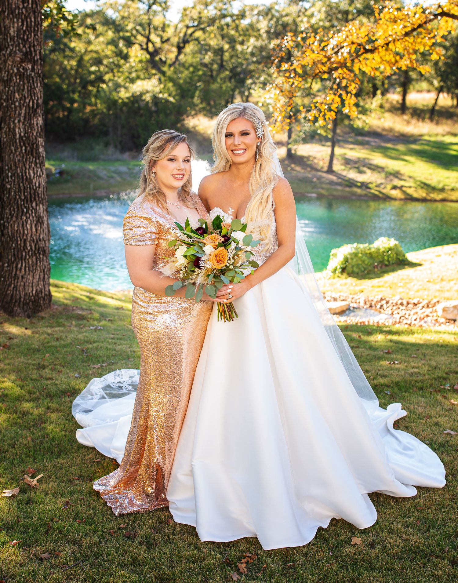 Wedding party members pose by a scenic pond with fall foliage in a beautiful outdoor setting.