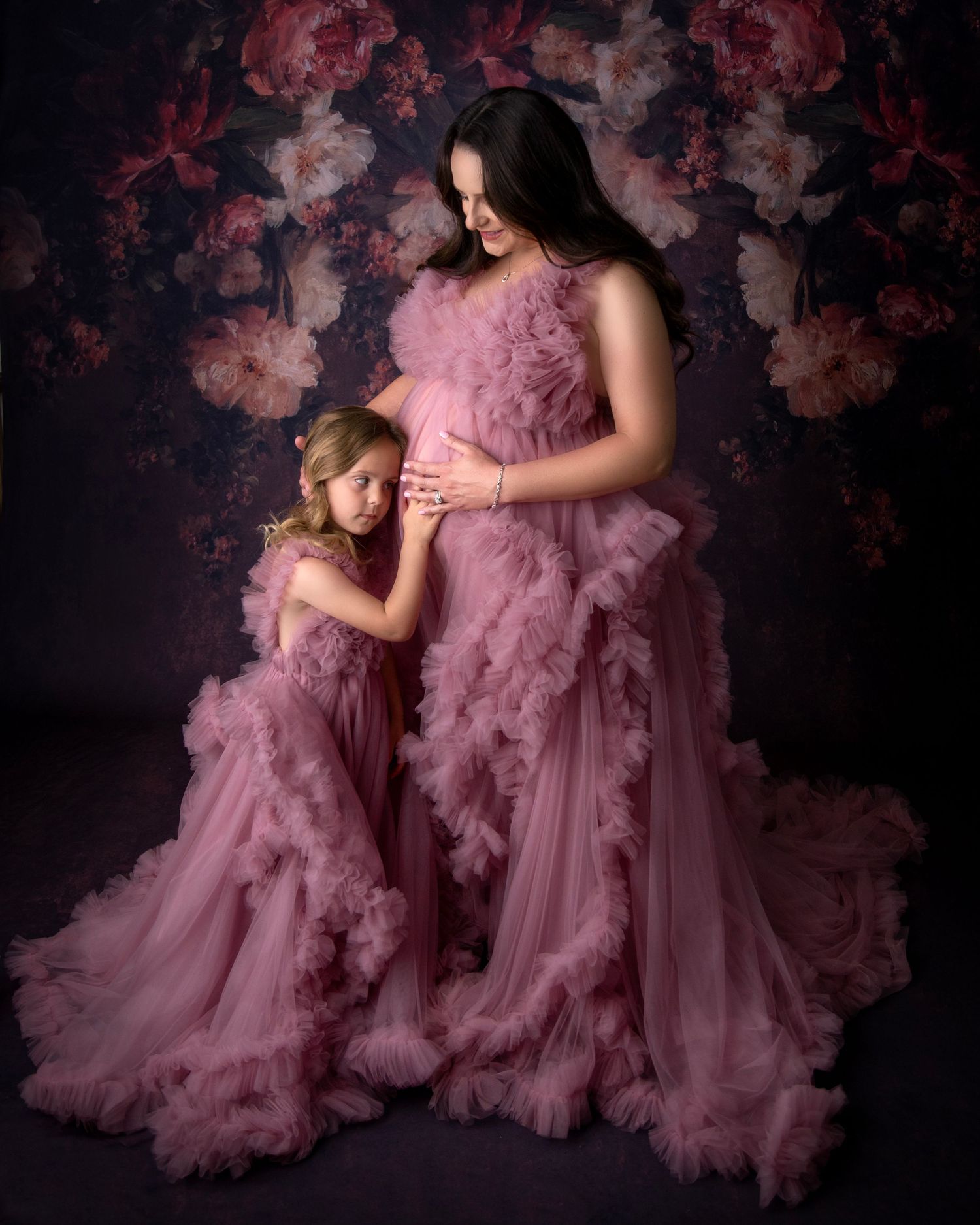Mother and daughter wearing matching pink ruffled tulle gowns pose against dark floral backdrop for maternity photo.