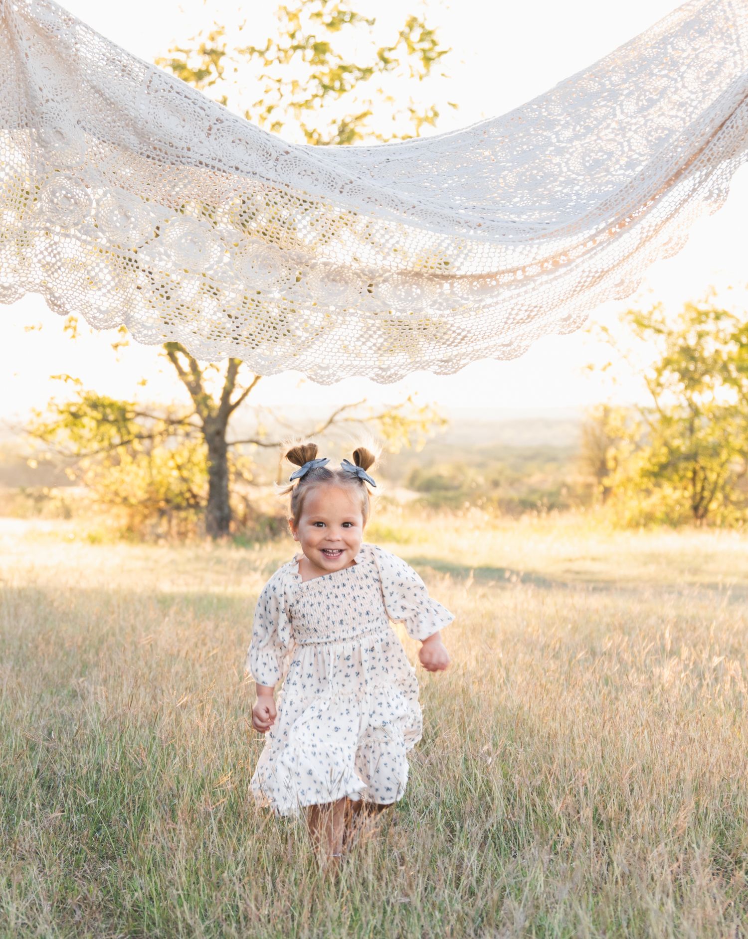 A toddler in a white patterned dress twirls joyfully in a golden sunlit field during sunset.