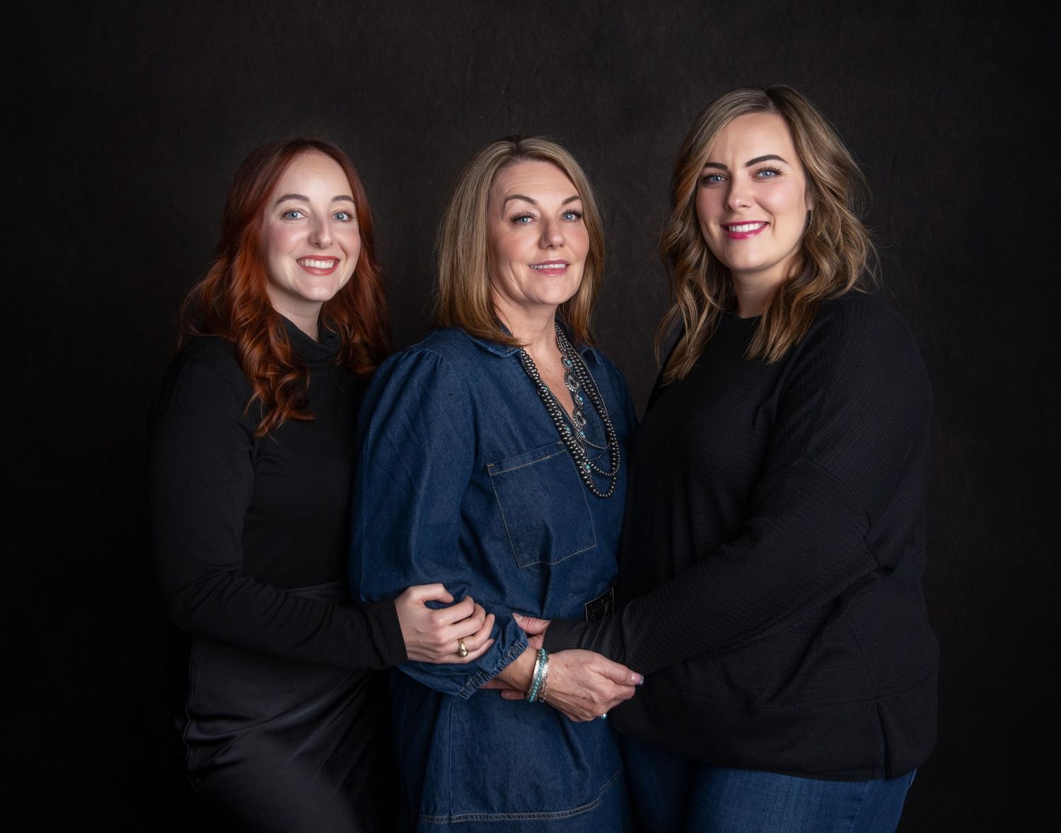 Three women in dark business attire pose together against a black background in a professional studio setting.