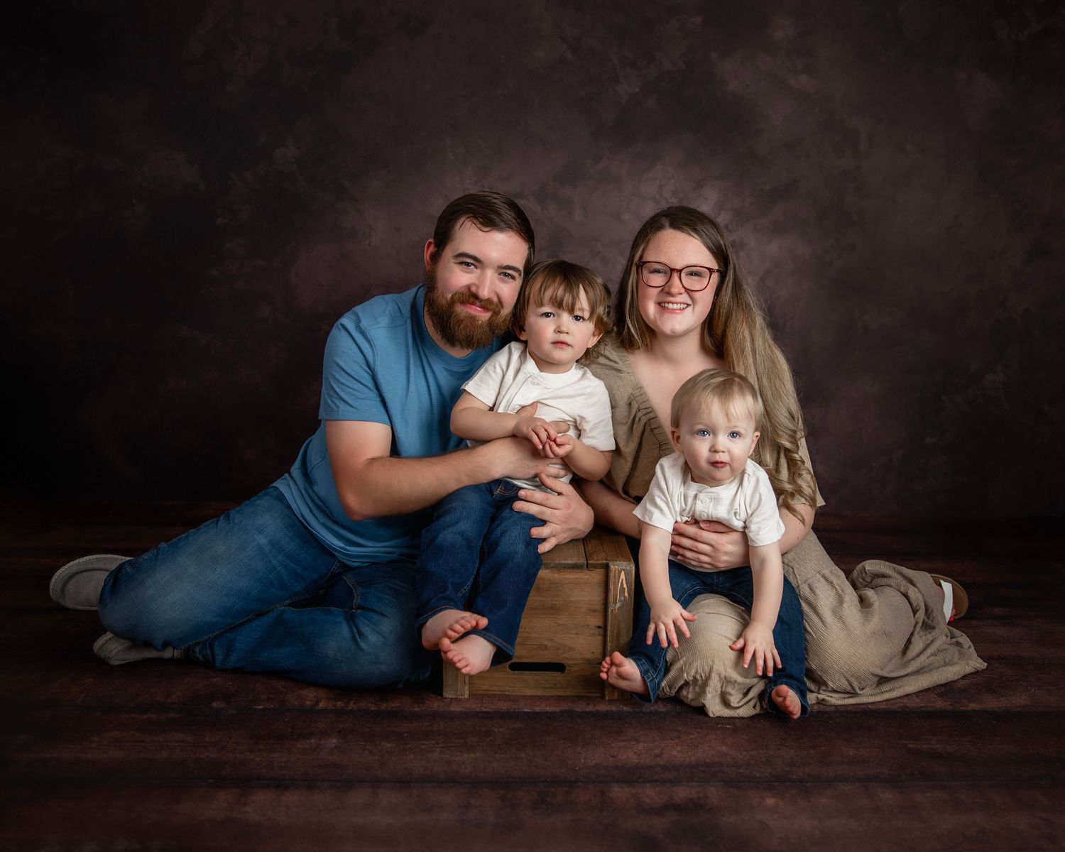 A studio portrait of a family sitting together on wooden furniture against a dark background.