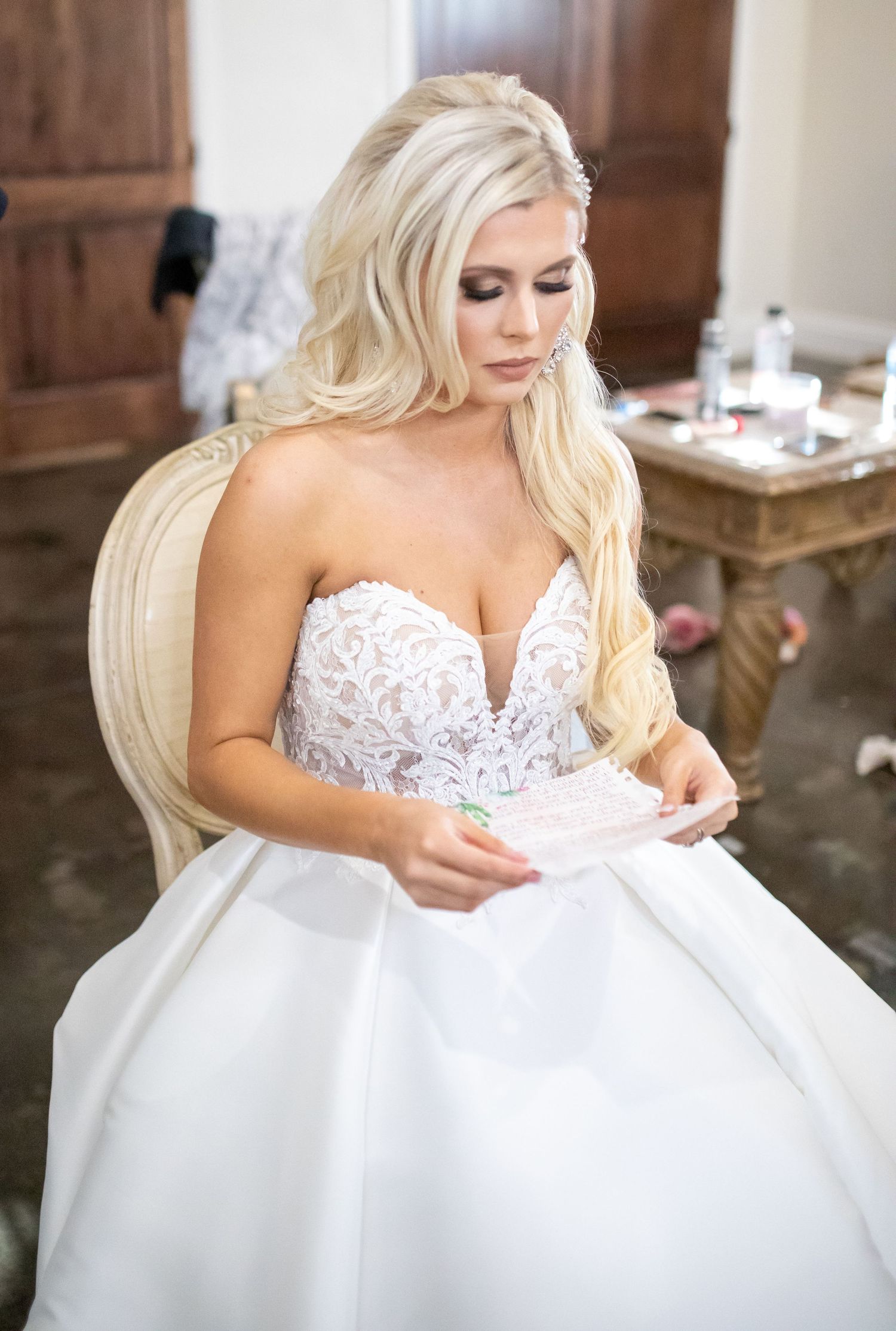A bride in a strapless lace wedding dress sits reading a note in an elegant getting-ready room.