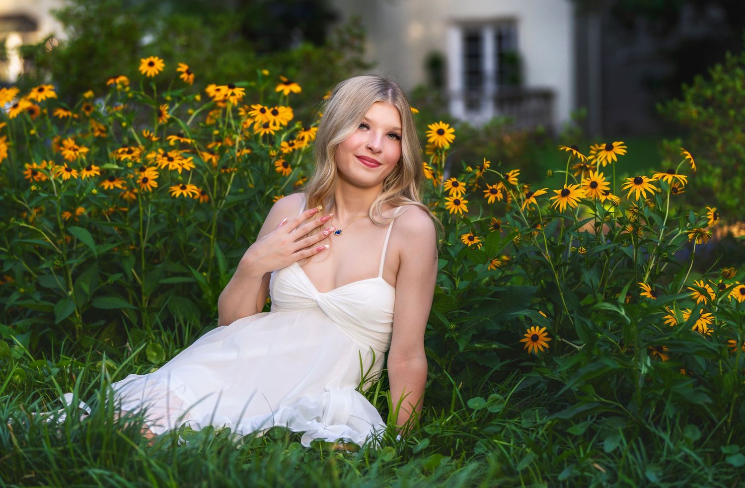 Young blonde woman in white dress sitting among vibrant yellow black-eyed Susan flowers in a lush garden.