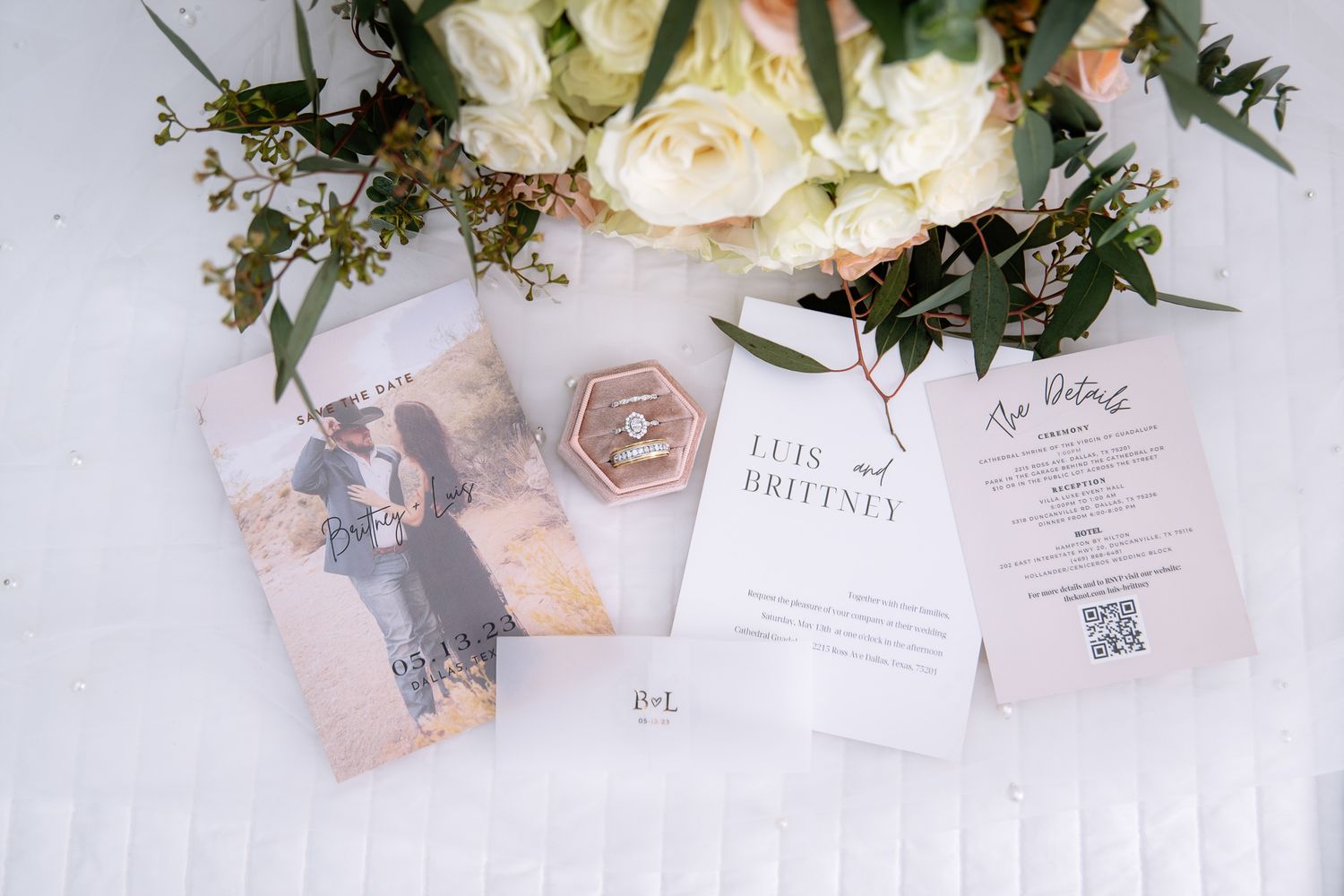 Wedding stationery and floral arrangements with white roses and greenery on a white tablecloth.