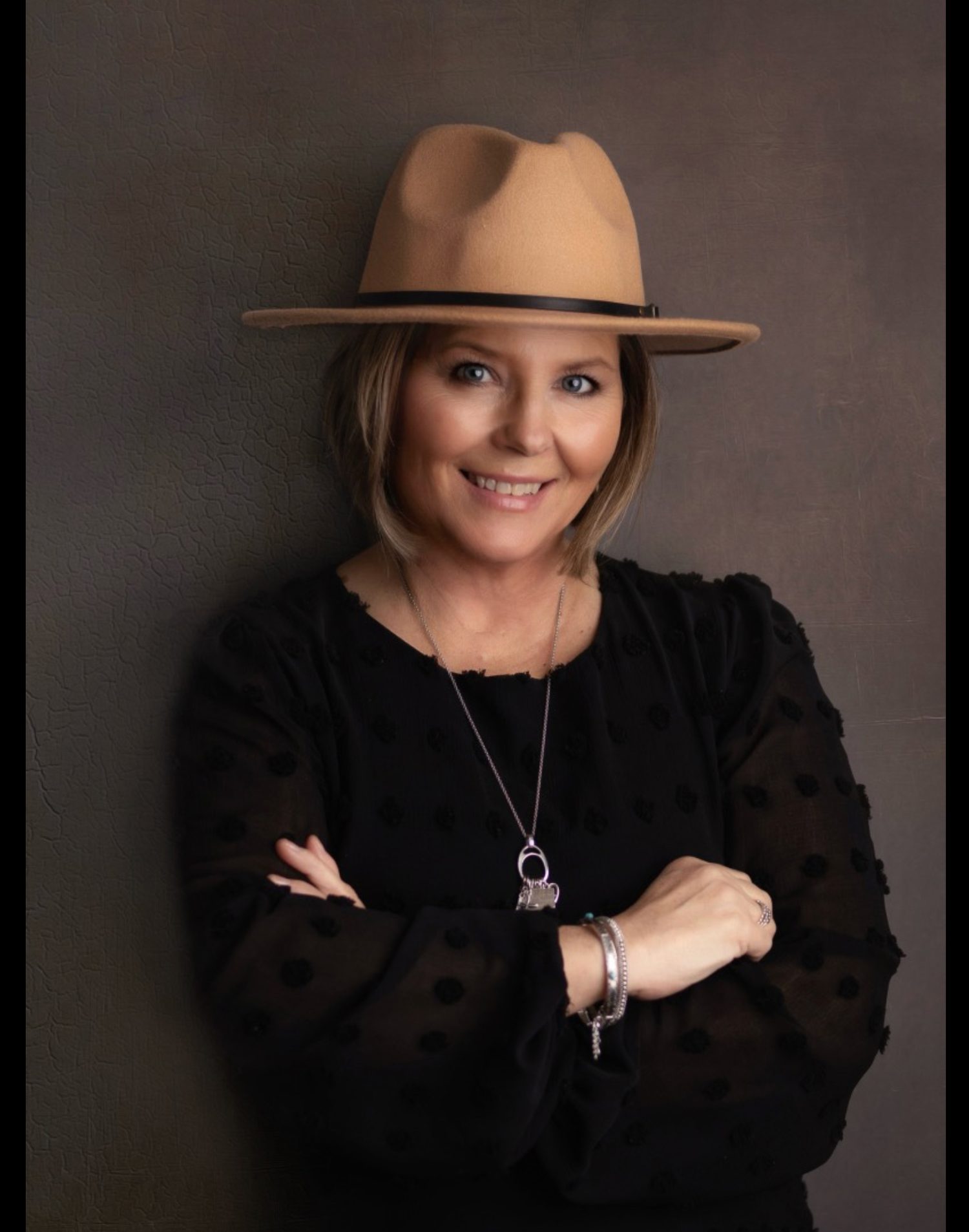 Professional portrait of woman in tan fedora and black blouse against dark background.