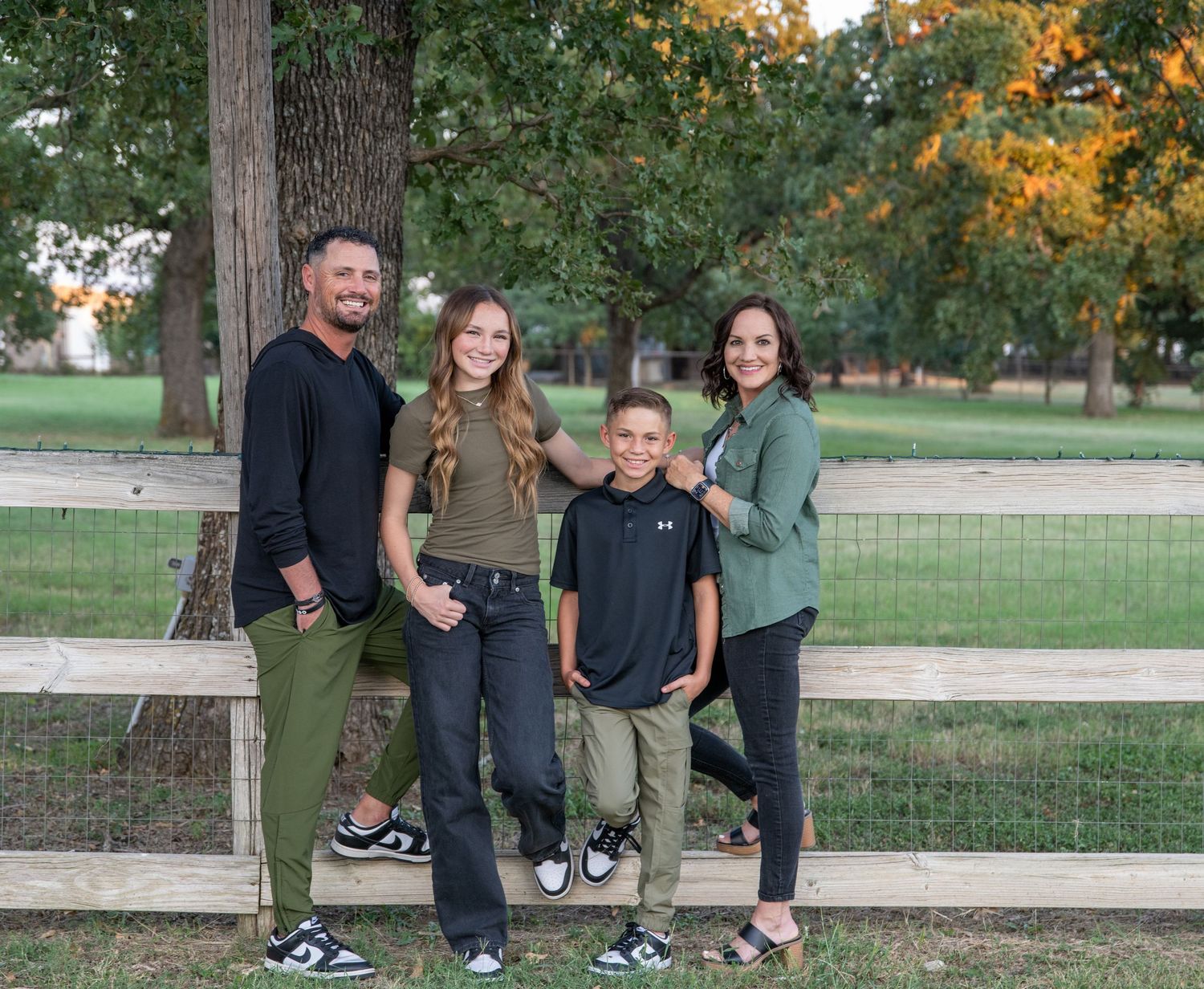 Family leaning on white wooden fence with autumn trees in background.