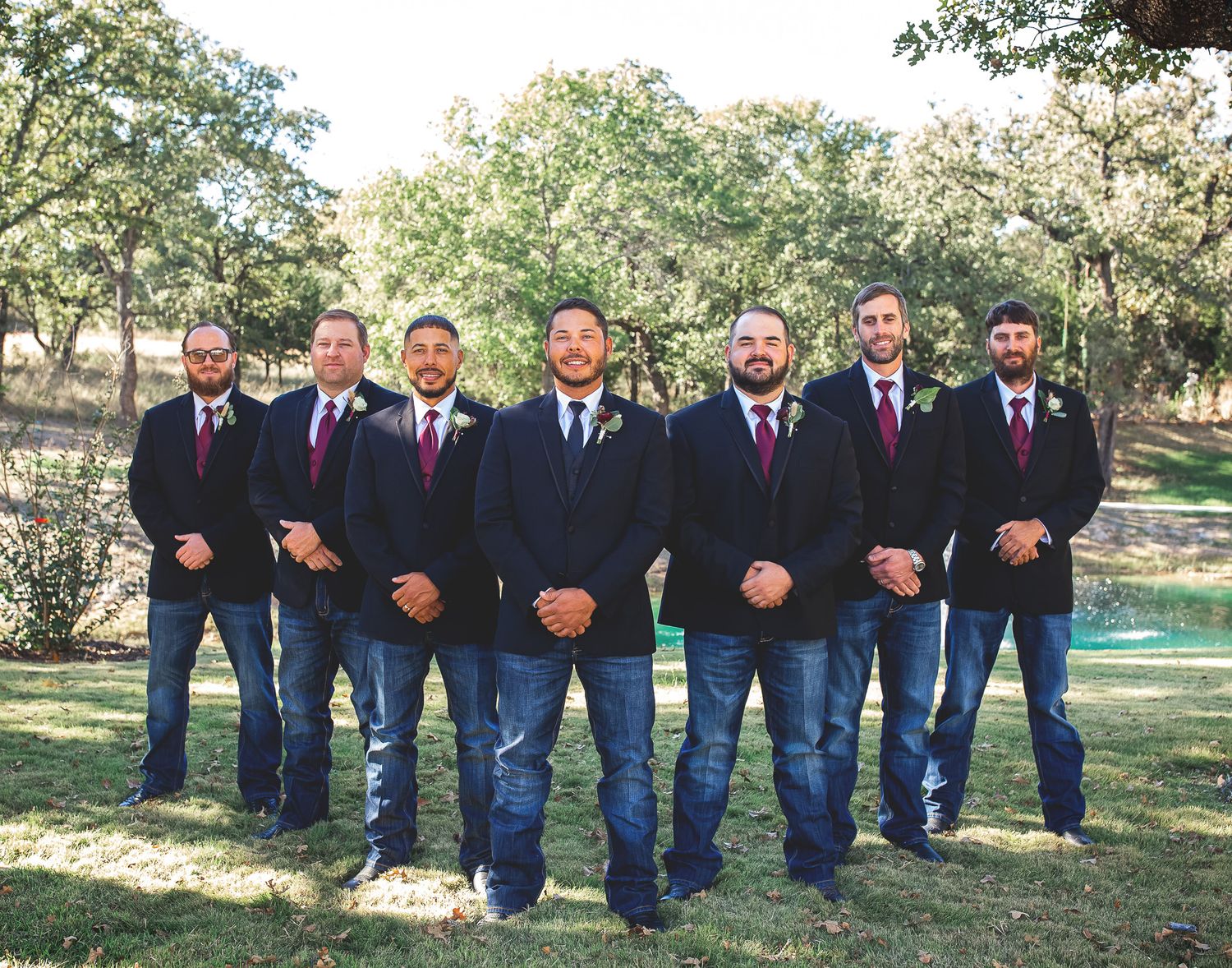 Groomsmen in matching blue jeans and suit jackets pose together outdoors against a wooded backdrop.