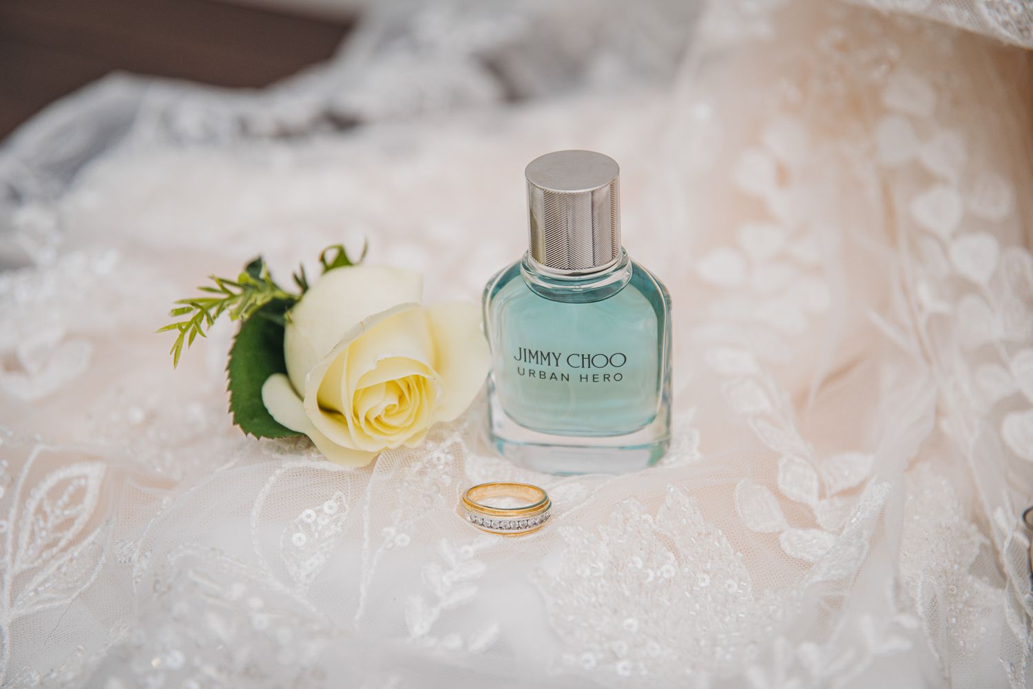 Wedding accessories including a blue perfume bottle, yellow rose, and gold ring on white lace.