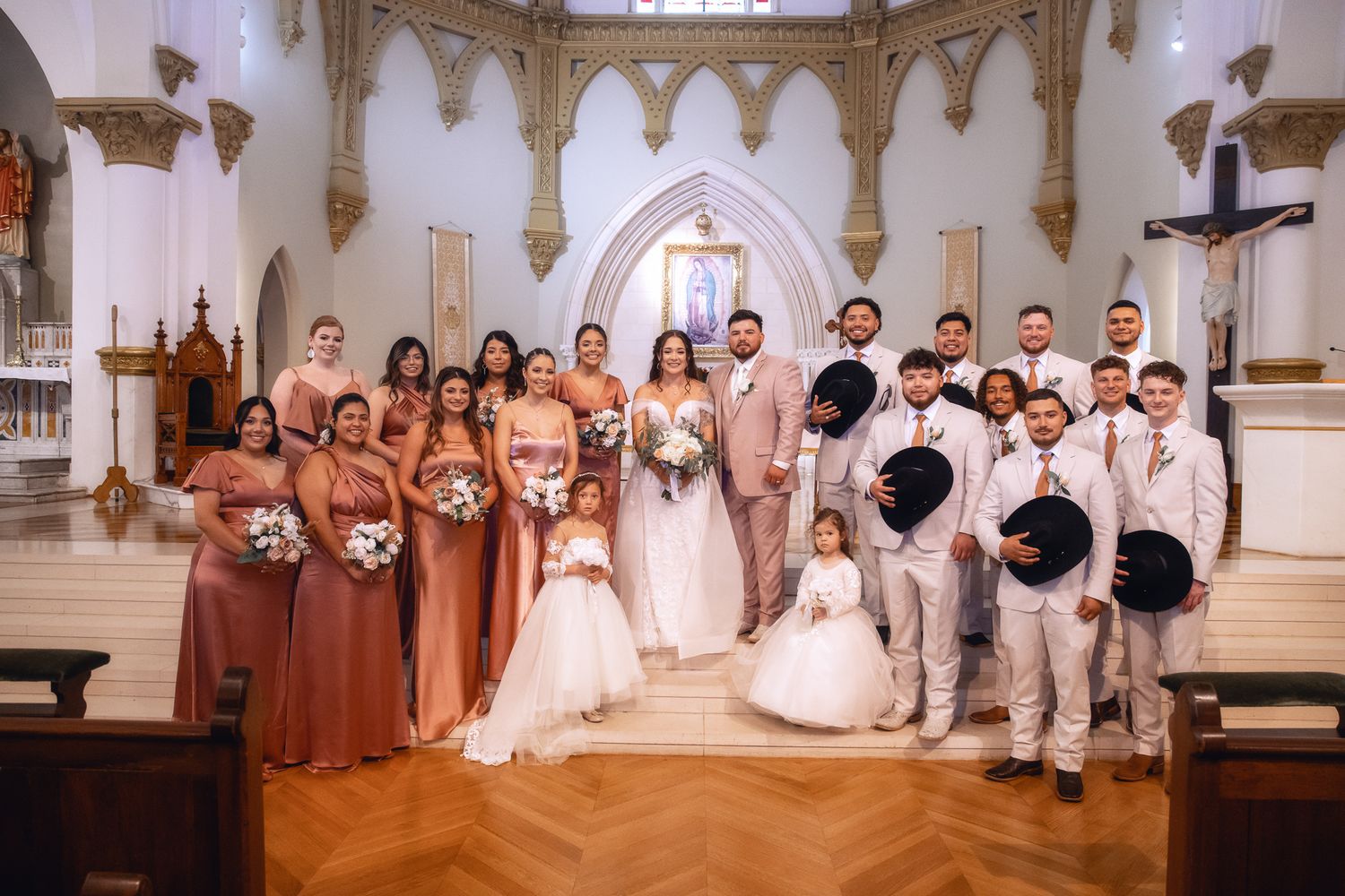 Wedding party poses together in church sanctuary with coral bridesmaid dresses and light suits.