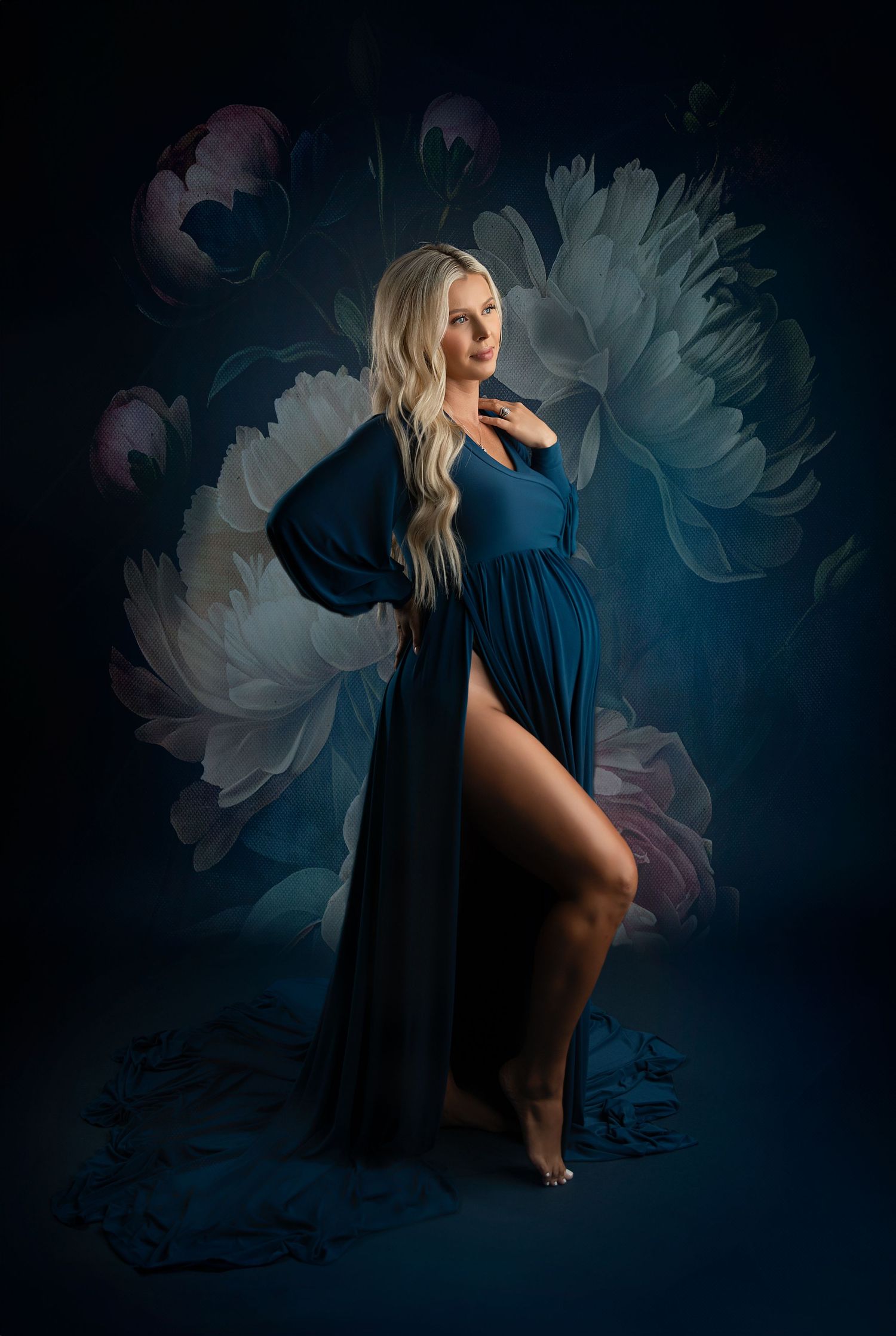Dramatic maternity portraits with flowing blue gown against dark background with floral elements.