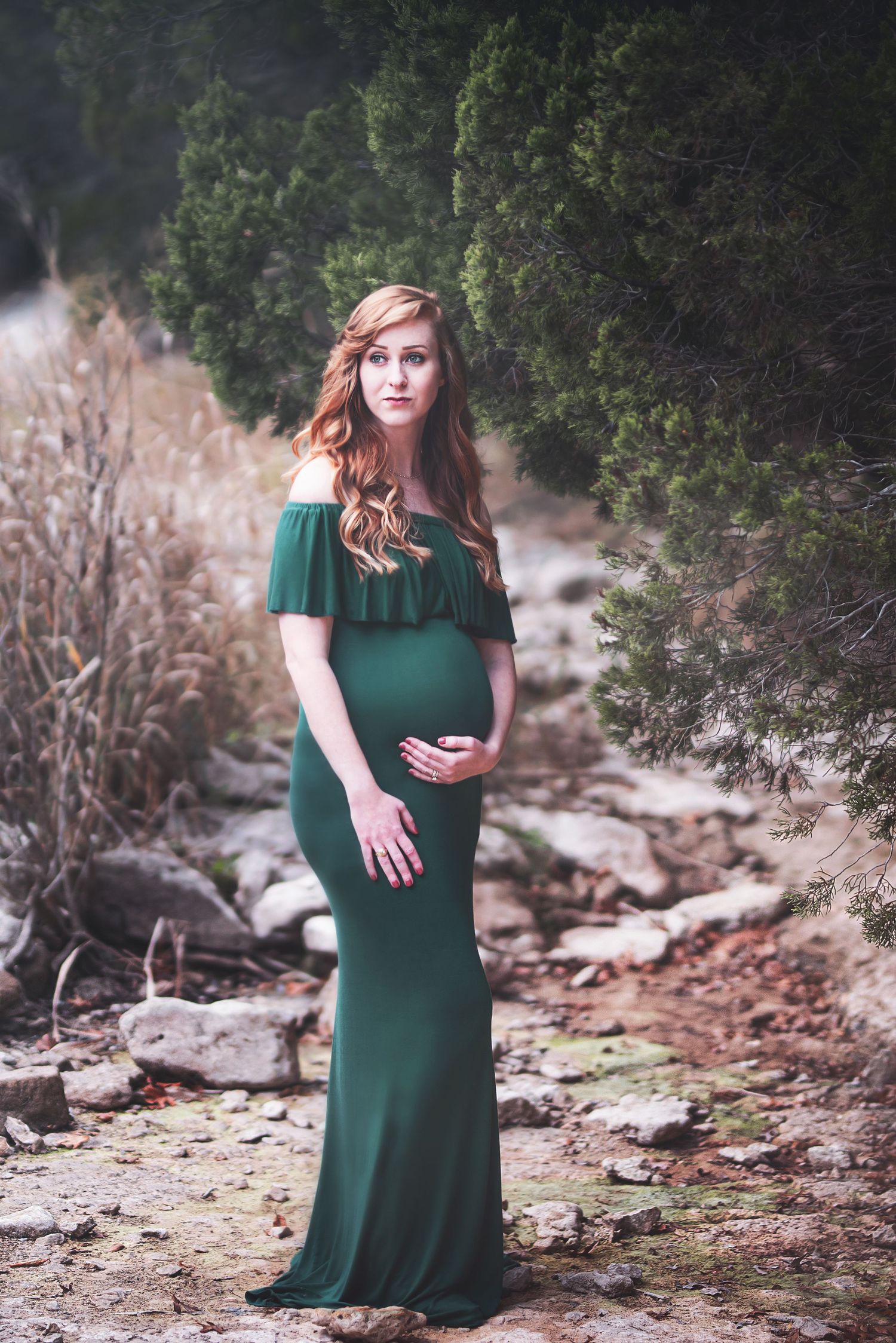 Woman in emerald green dress poses on rocky terrain during outdoor maternity session.
