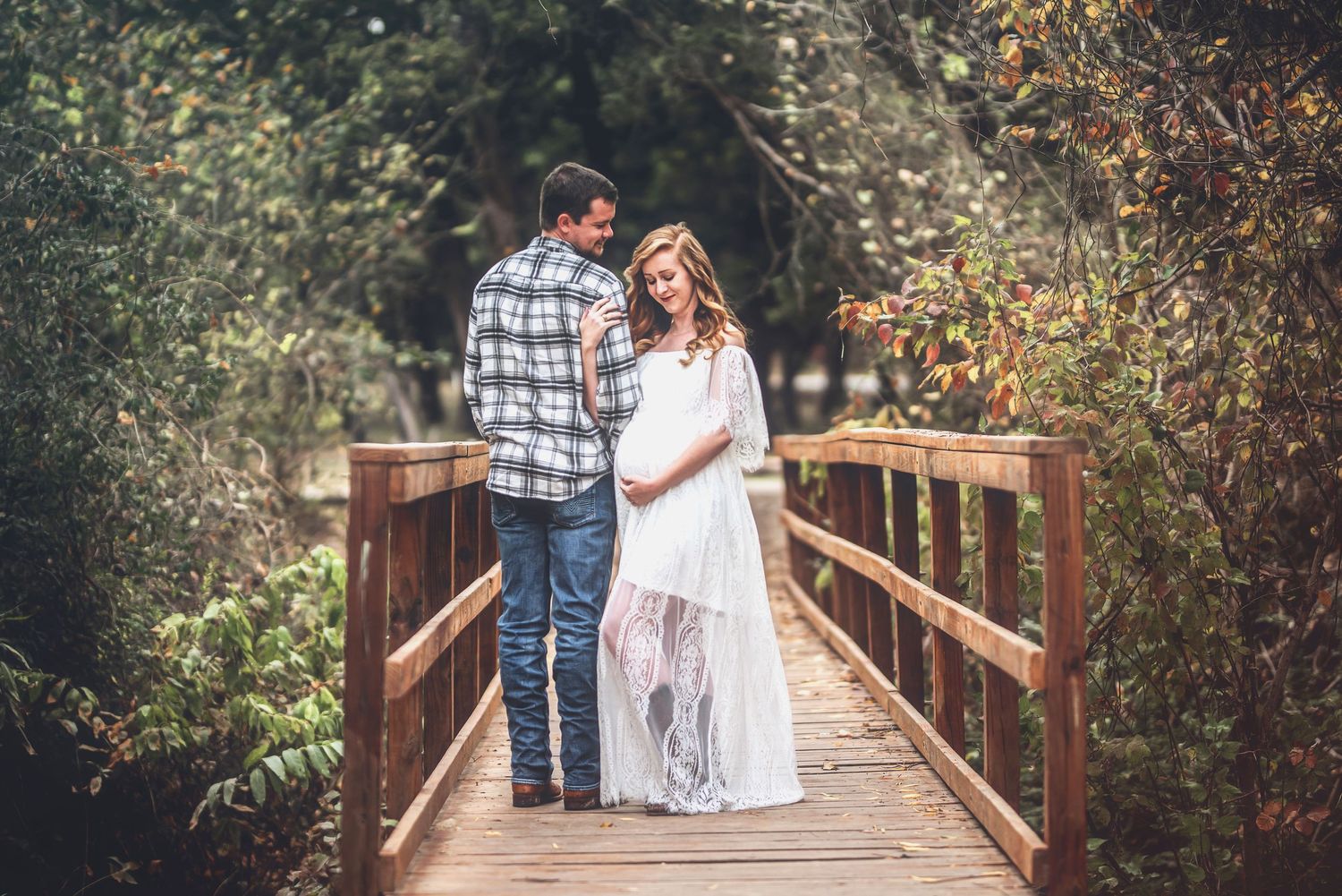 Expectant couple shares intimate moment on wooden bridge surrounded by lush greenery and autumn foliage.
