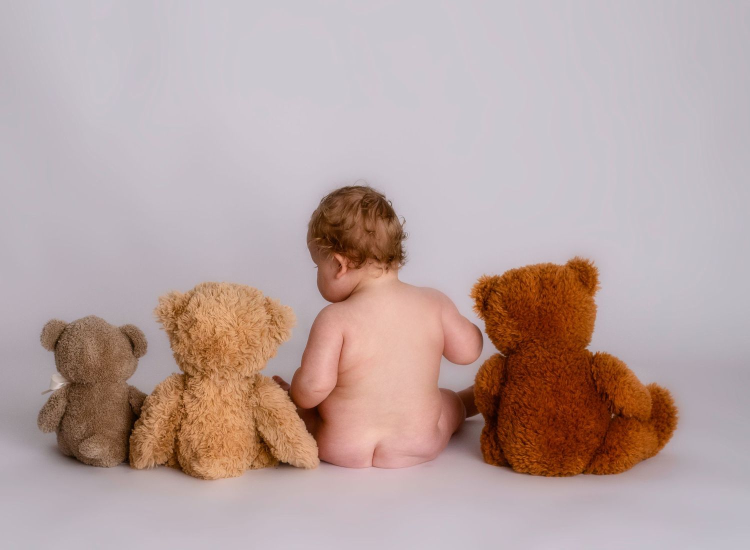 Baby sitting with three teddy bears in a simple studio setting against white background.