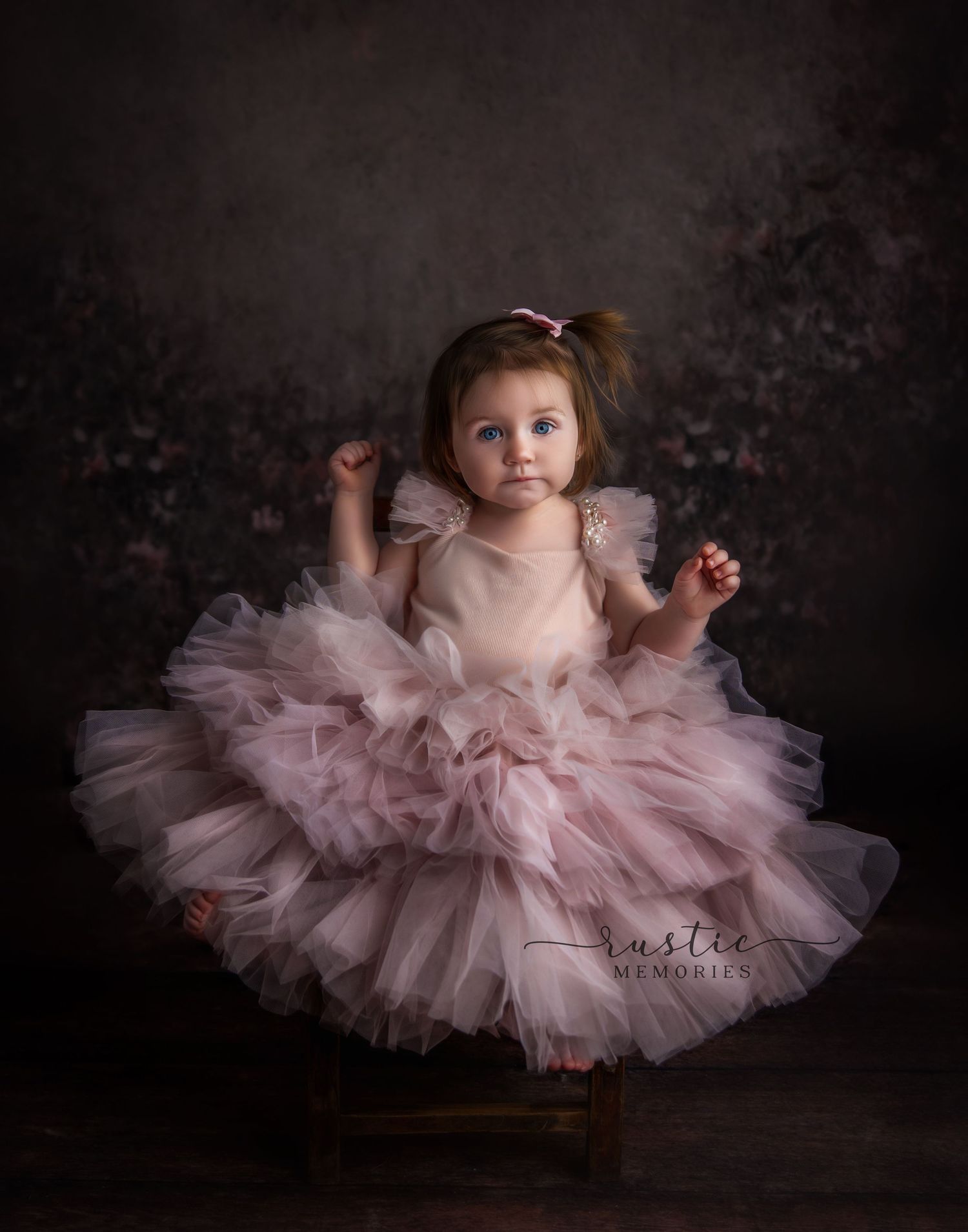 A baby wearing a fluffy pink tulle dress poses for artistic portraits against a dark backdrop.